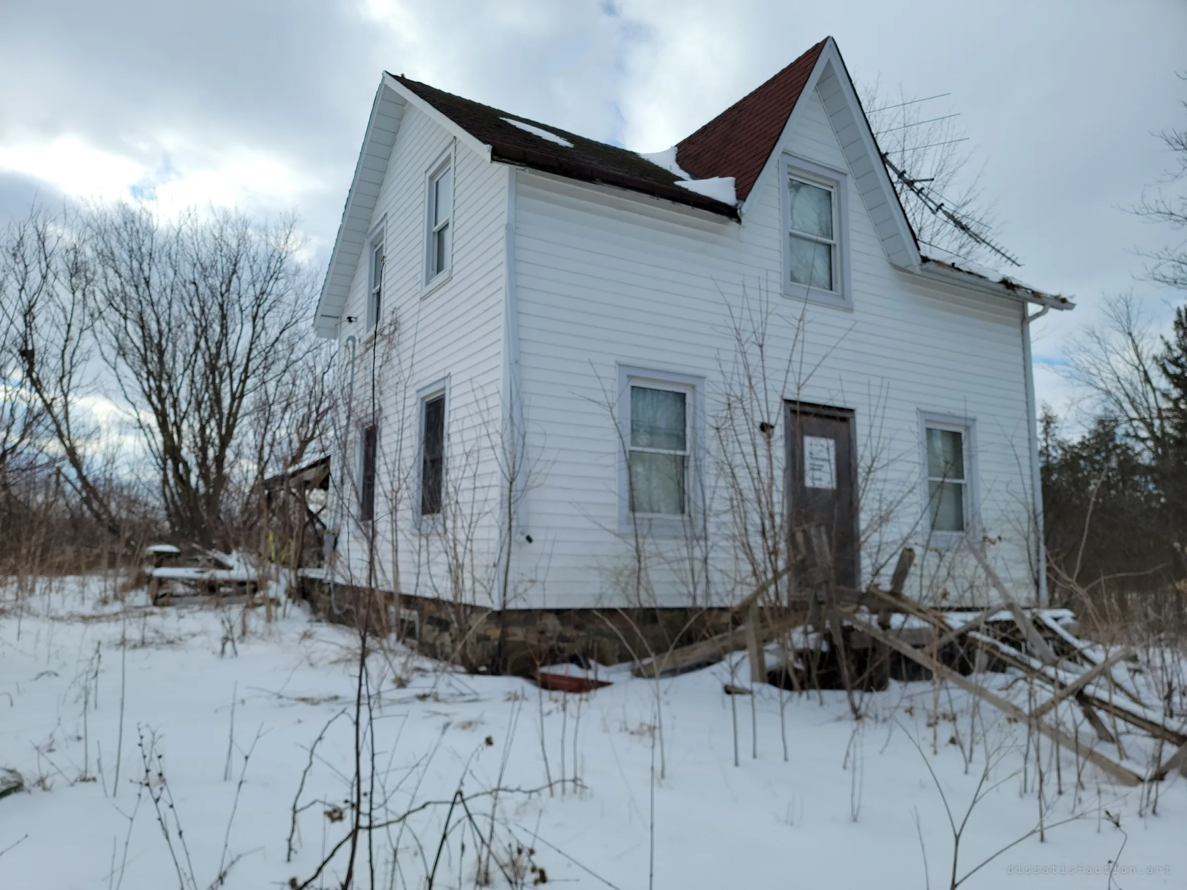 A white-sided farmhouse with a steep red-shingled gable stands in deep snow, its boarded door posted with a faded notice and a collapsed fence leaning against the wall. The bent antenna and missing shingles show years without anyone living here.