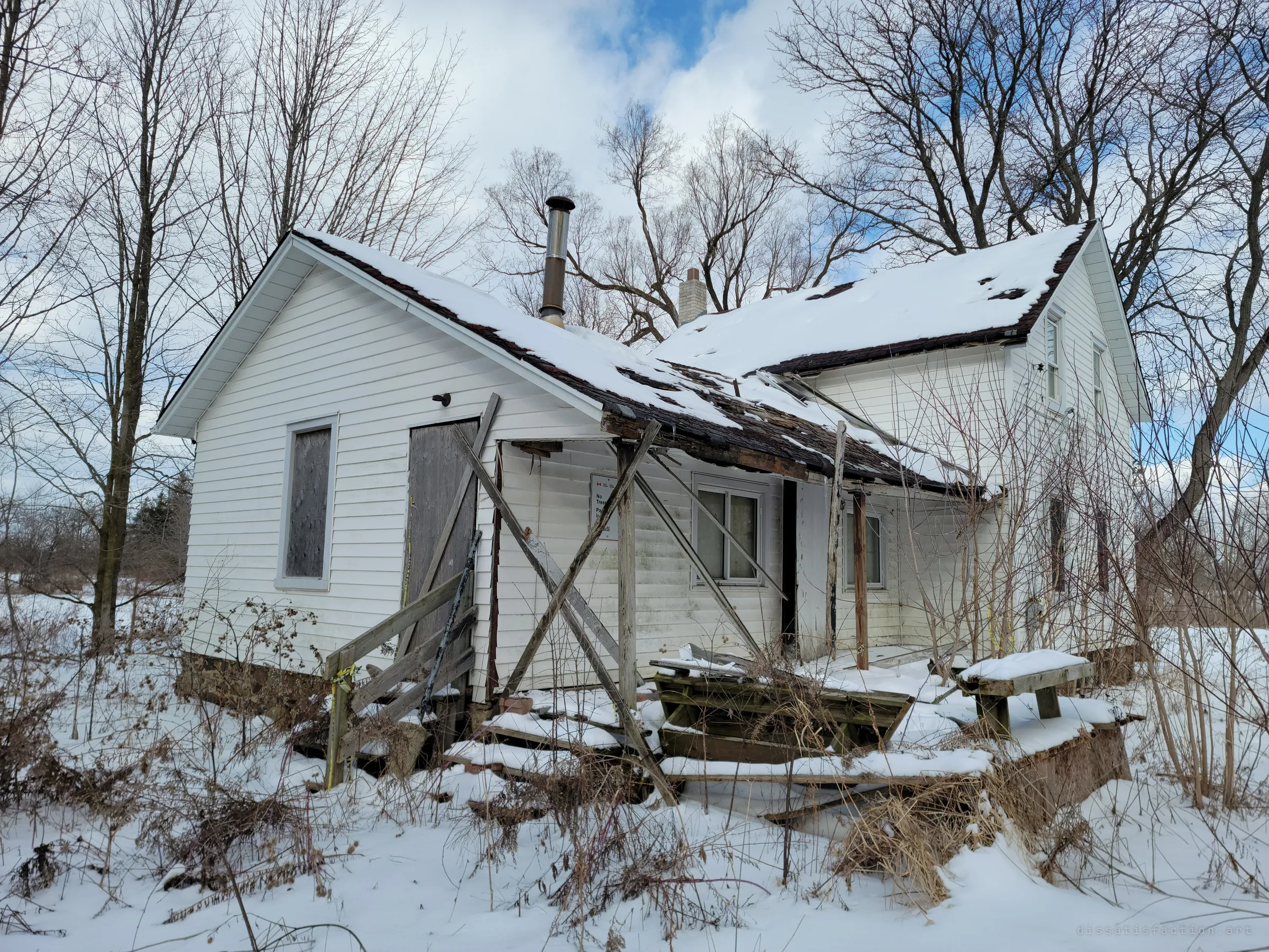 The back of the house under a snow-loaded roof: the porch roof has caved in and is propped with crossed timbers, shingles peeling away above a boarded door. Two chimneys still rise through the snow among bare trees.