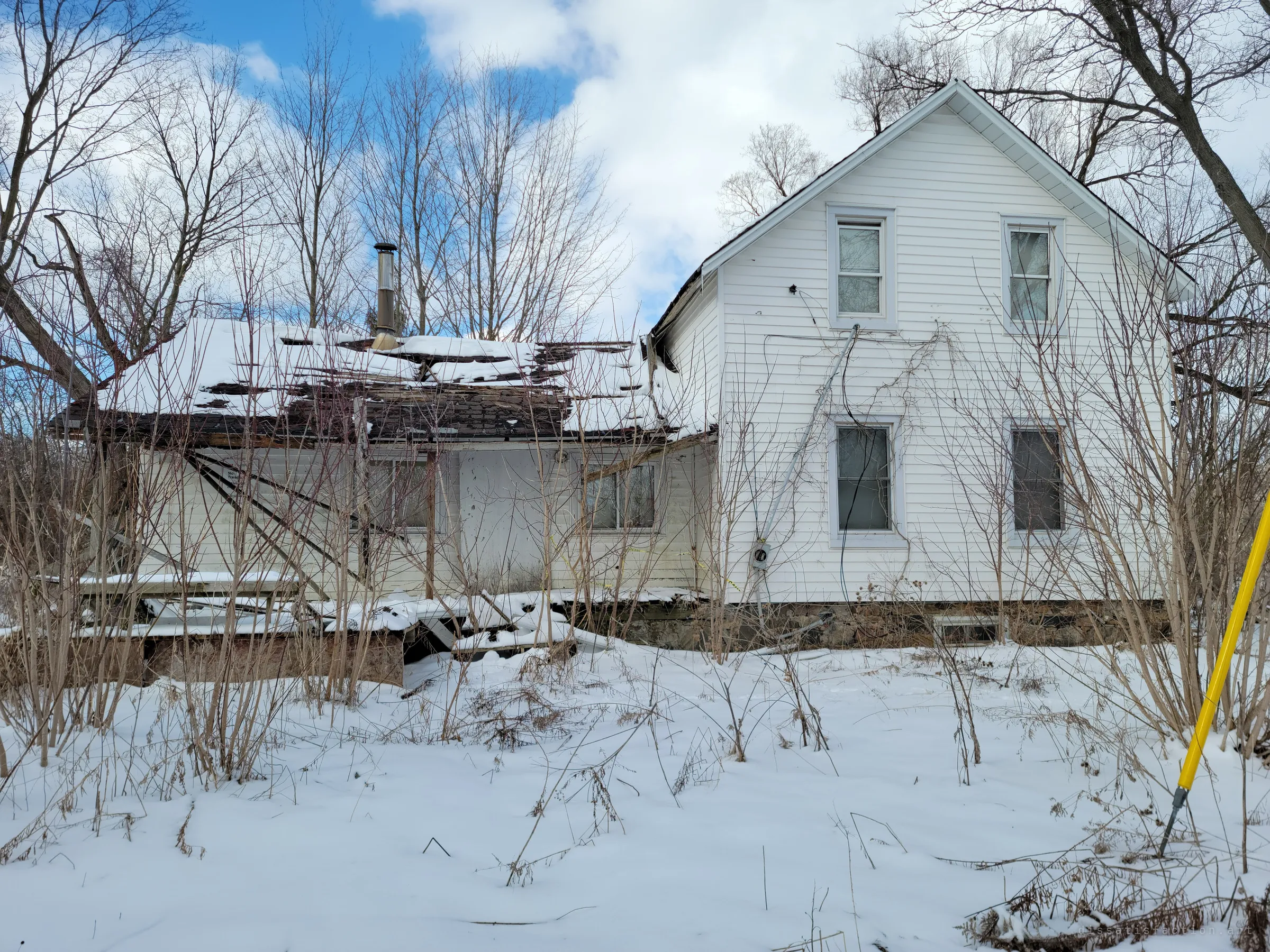 Side view of the house with its rear addition collapsing, the porch roof sagging into a tangle of broken boards and snow. Bare saplings have grown up against the siding, and a yellow marker stake stands in the foreground.