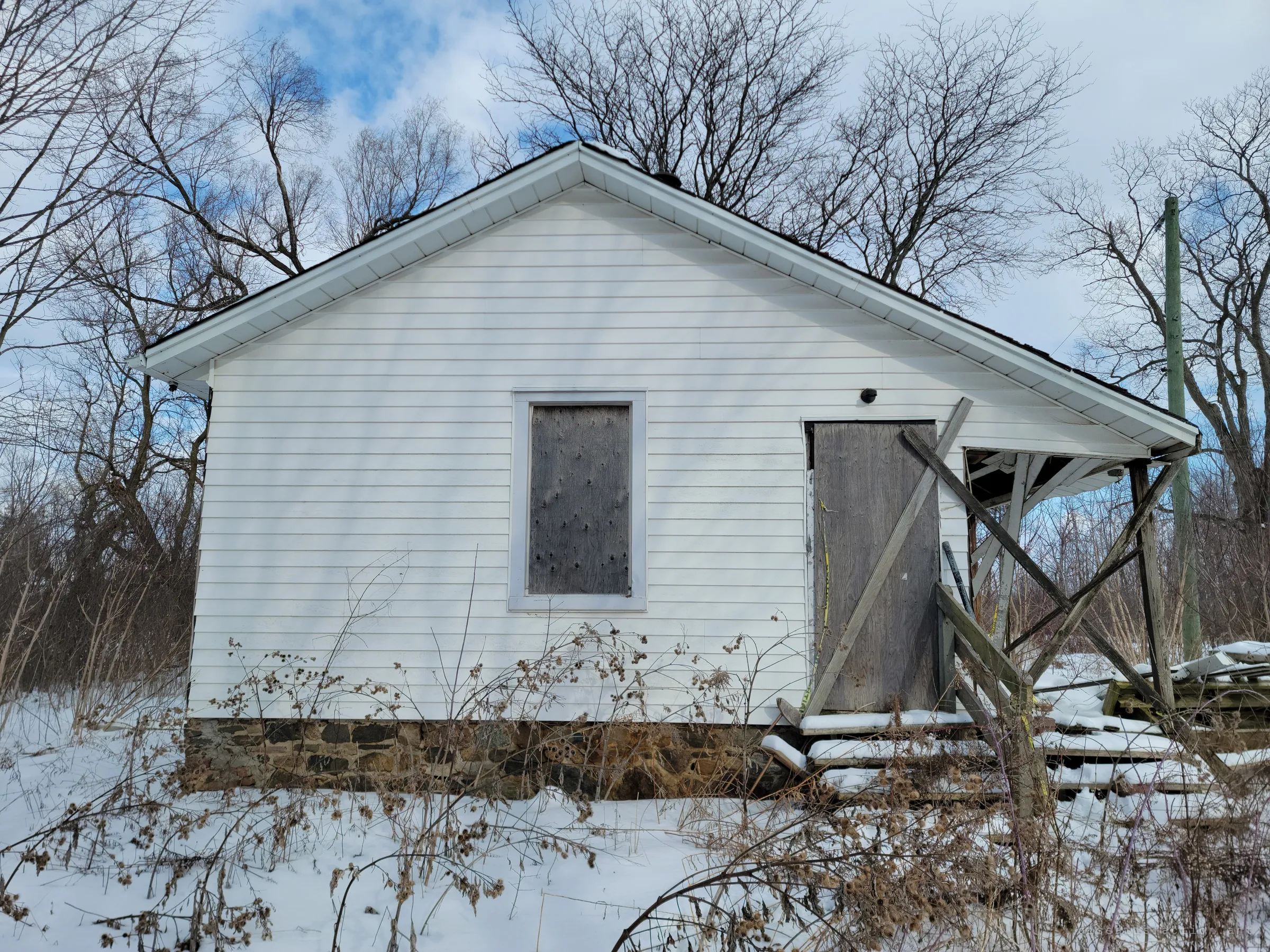 The gable end of the rear addition, its window and door sealed with weathered plywood above a fieldstone foundation. A broken porch frame leans off the corner, and burdock stalks crowd the snow in front.