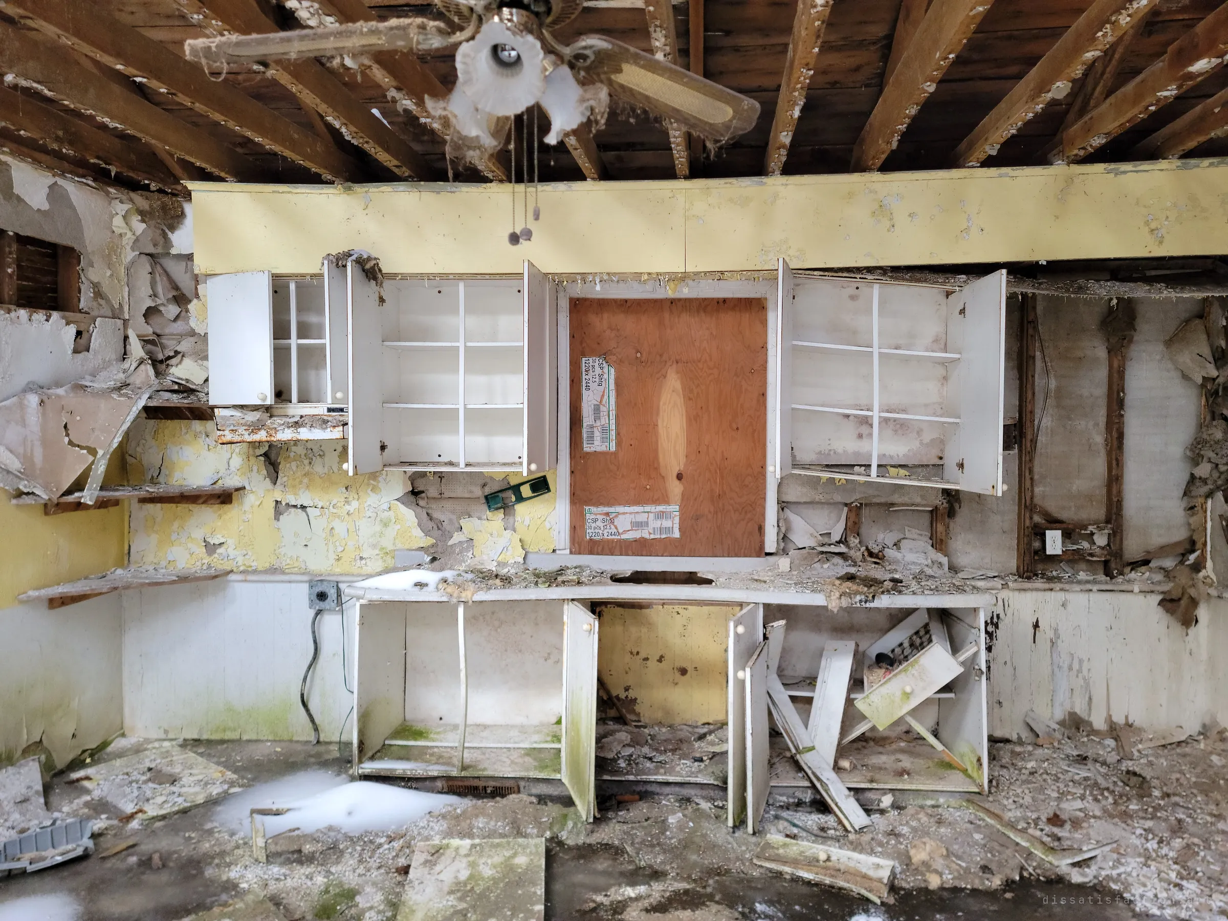 A gutted kitchen with open, empty cabinets hanging under exposed joists and a dusty ceiling fan, the window behind them boarded with stamped plywood. Peeling yellow paint, torn drywall and snow drifted onto the floor show how open the house is to the weather.