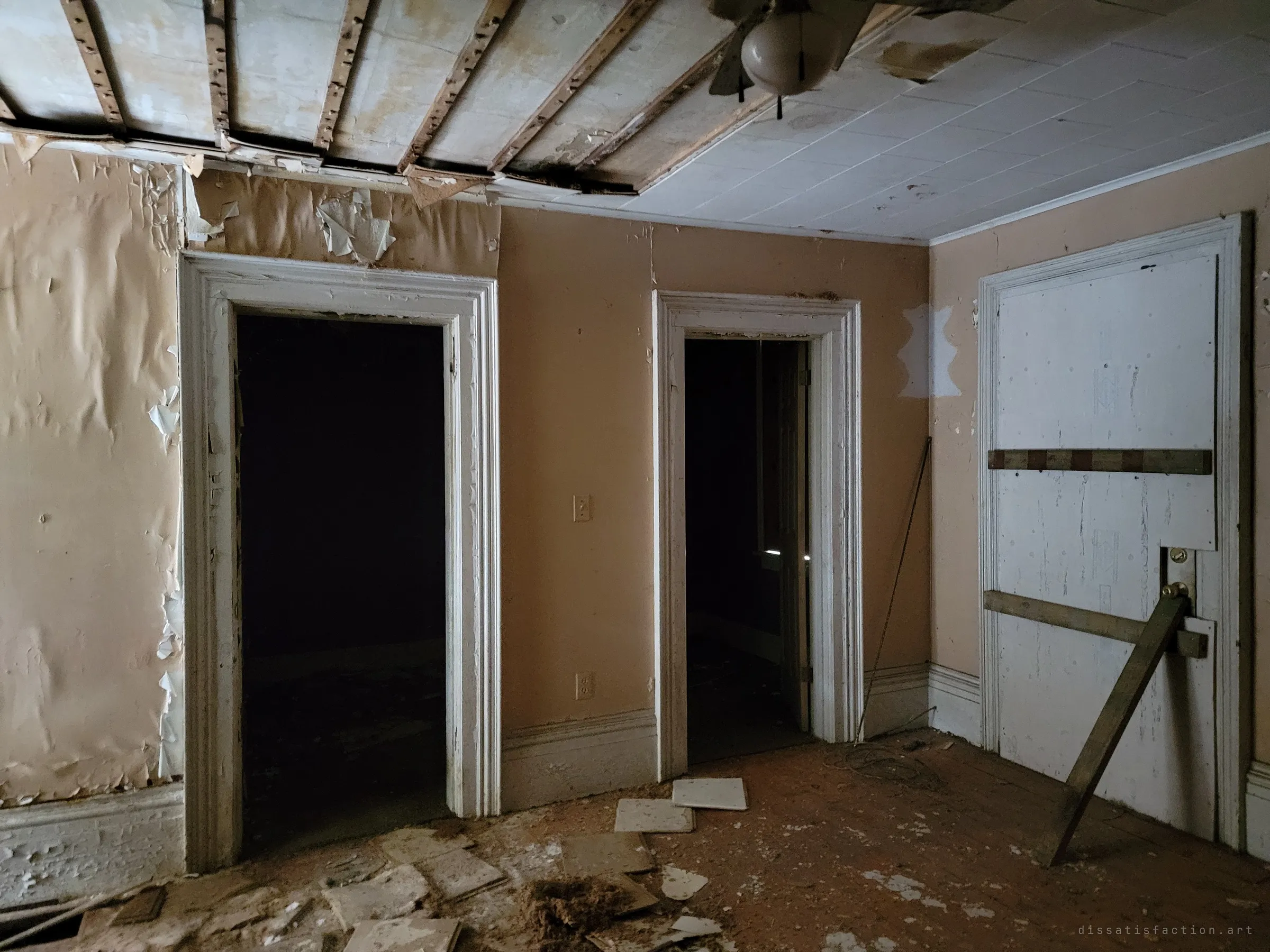 A dim room with peach-painted walls and heavy white door trim, two doorways opening into darkness and an outside door boarded and braced with a timber. Ceiling boards hang loose and broken tiles litter the wooden floor.
