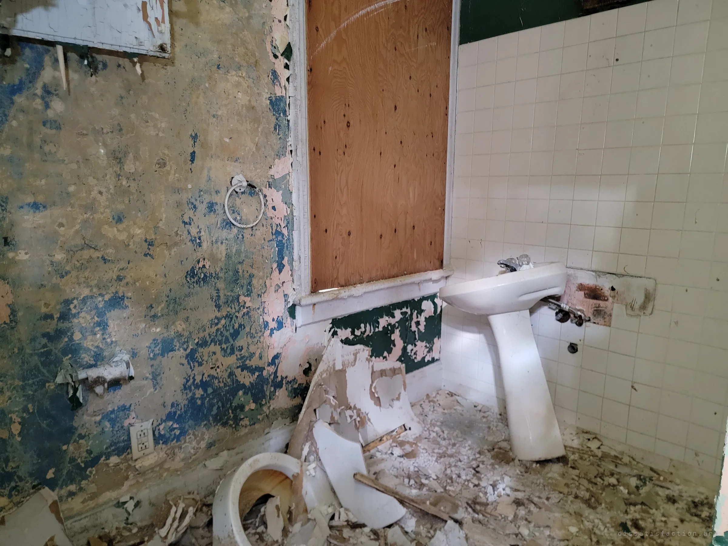 A wrecked bathroom where blue and green paint peels off the plaster around a boarded window. The toilet lies smashed on the floor beside a pedestal sink still clinging to the tiled wall.