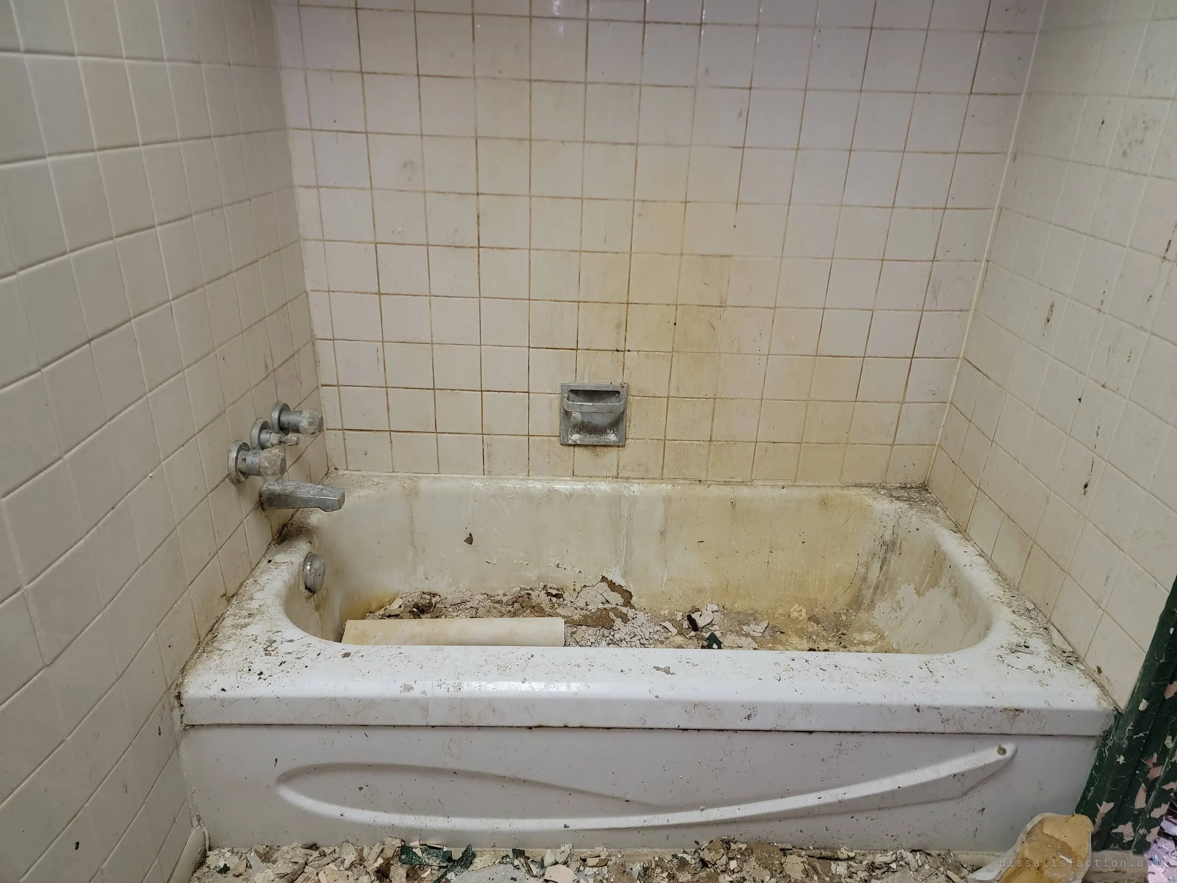 A grimy tiled bathroom with a stained white tub half full of fallen plaster and a length of pipe. Rust streaks and debris show how far the house has gone.