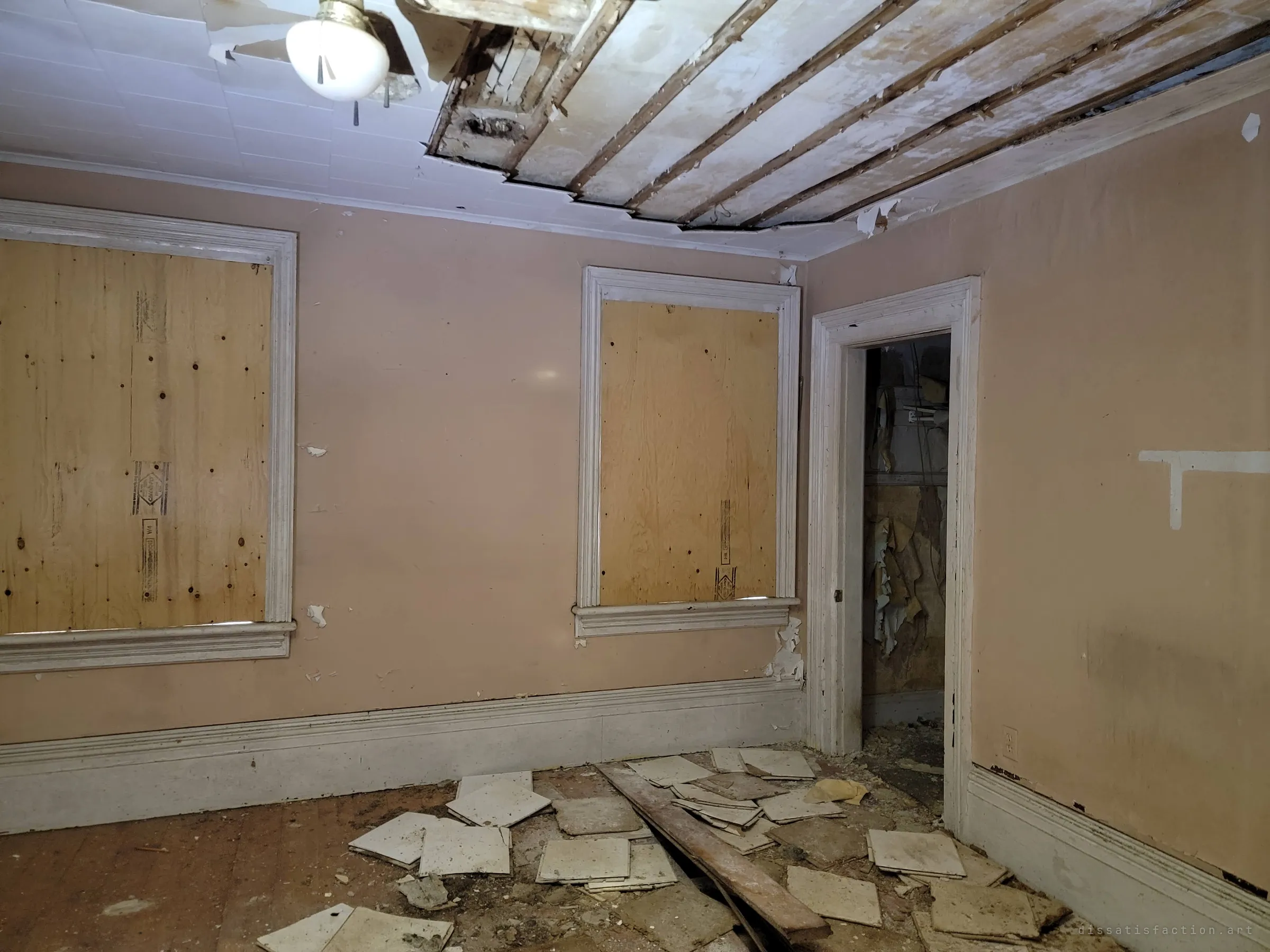 A peach-painted room with plywood over both windows, a ceiling torn open to the lath and old ceiling tiles scattered across the floor. The boarded windows mark the house as sealed off.