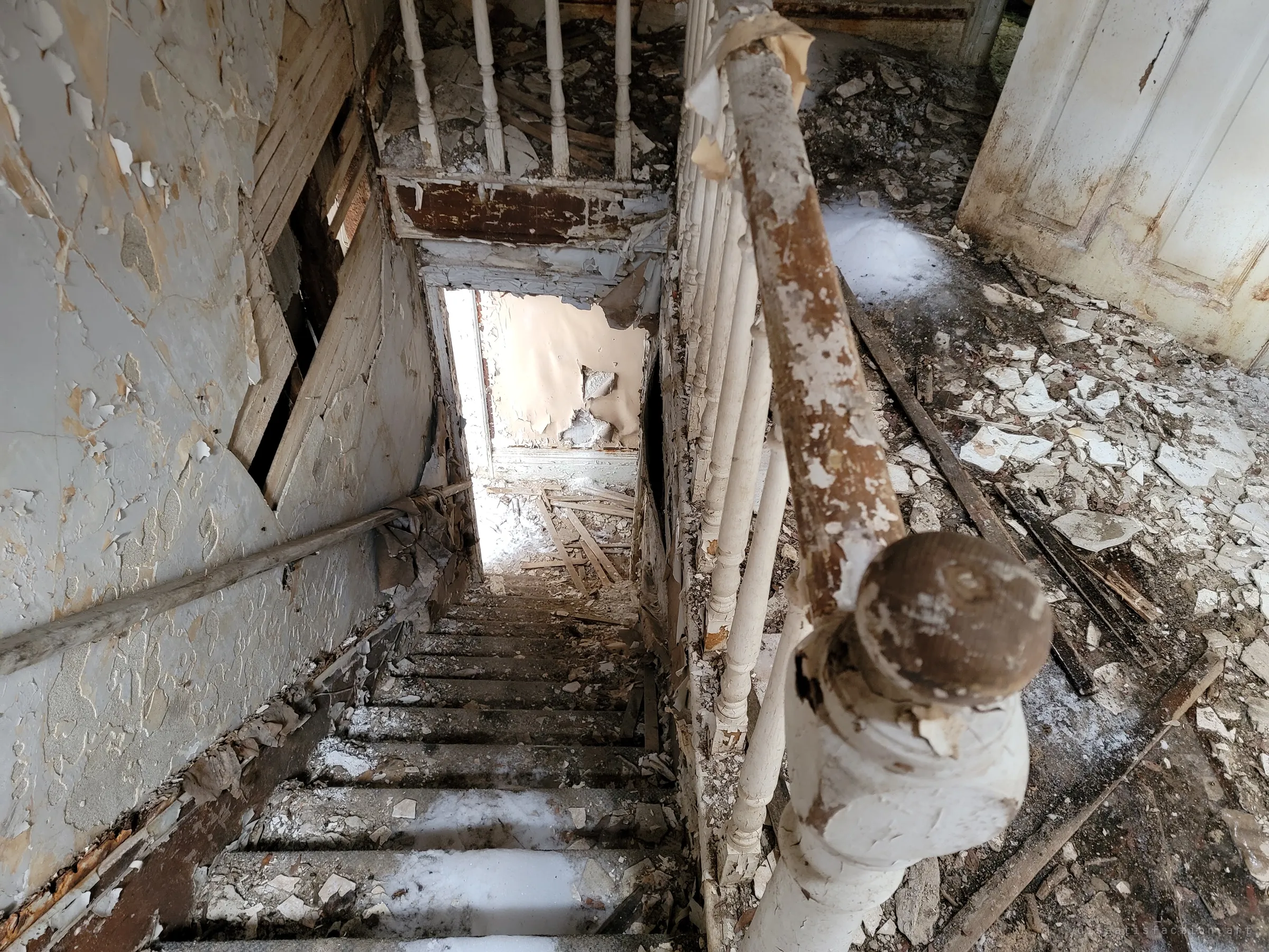Looking down a narrow staircase past a flaking newel post and banister, with snow drifted onto the steps and landing. Paint peels from every surface, showing the building open to the weather.