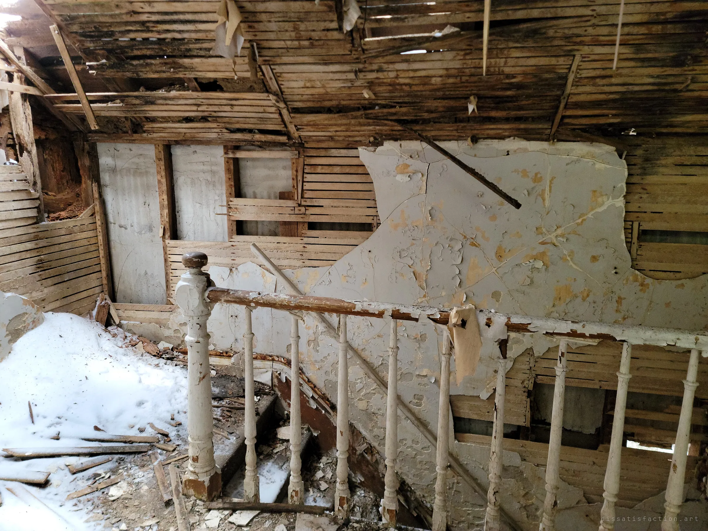 An upstairs landing where the ceiling and walls are stripped to bare lath above a white spindle railing, with snow piled on the floor. The collapse lets winter straight into the house.