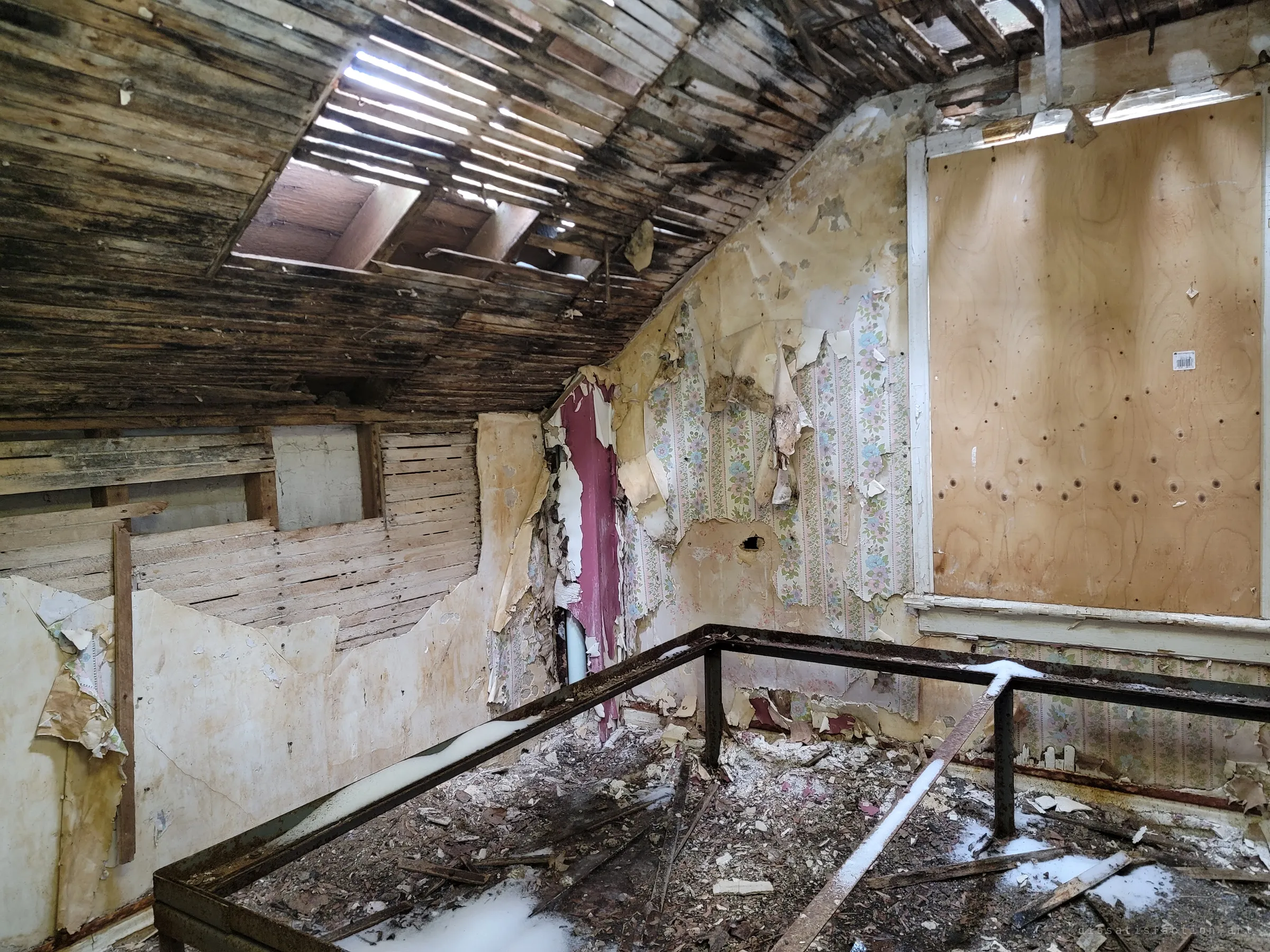 An attic bedroom under a sloped, rotting lath ceiling with daylight breaking through, floral wallpaper peeling away and a rusted bed frame dusted with snow. The open roof shows how far the decay has spread.