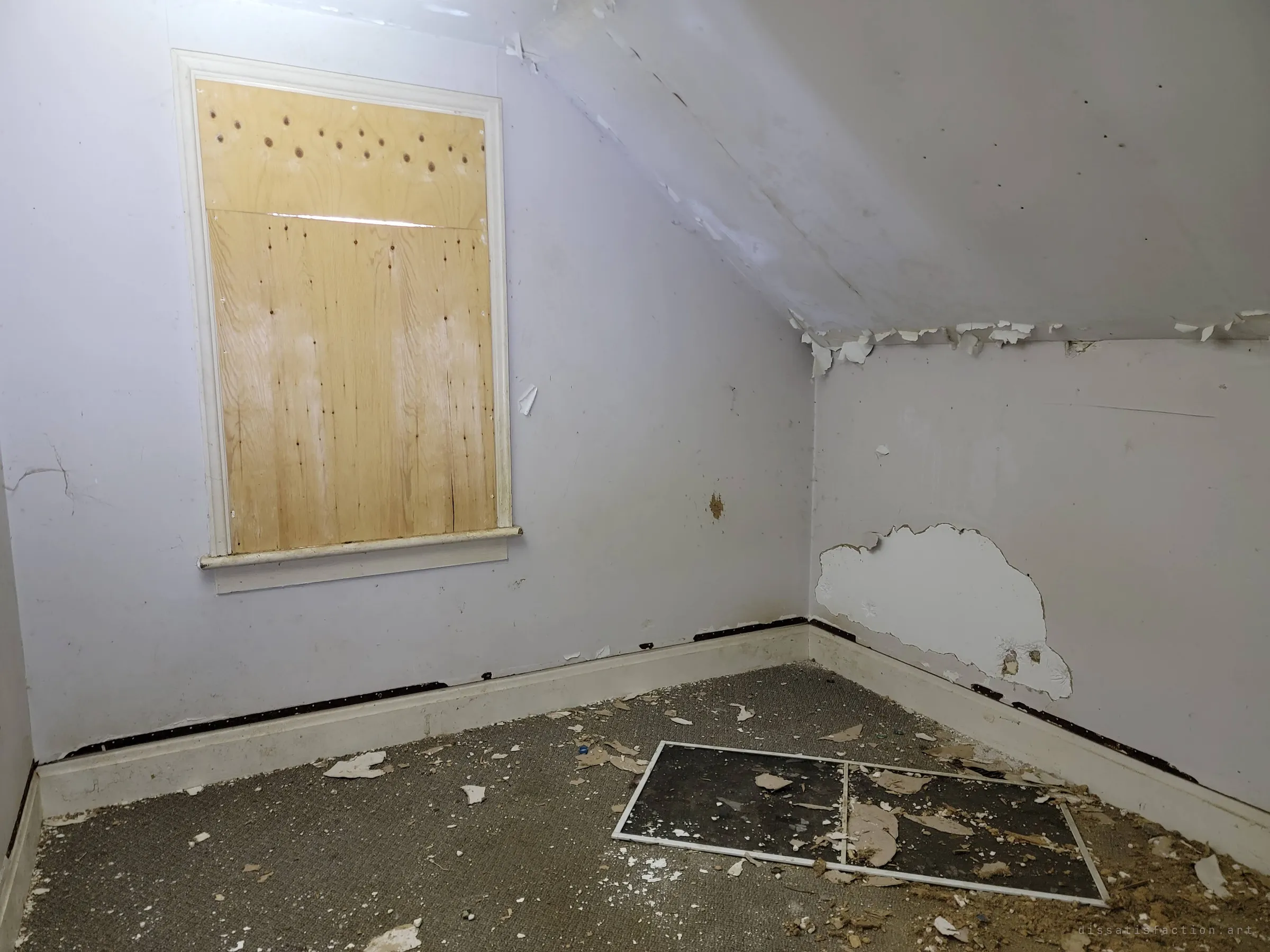 A small, pale attic room with a sloped ceiling, a plywood-covered window and a loose window screen lying on the dirty carpet. It is one of the few rooms left mostly intact.