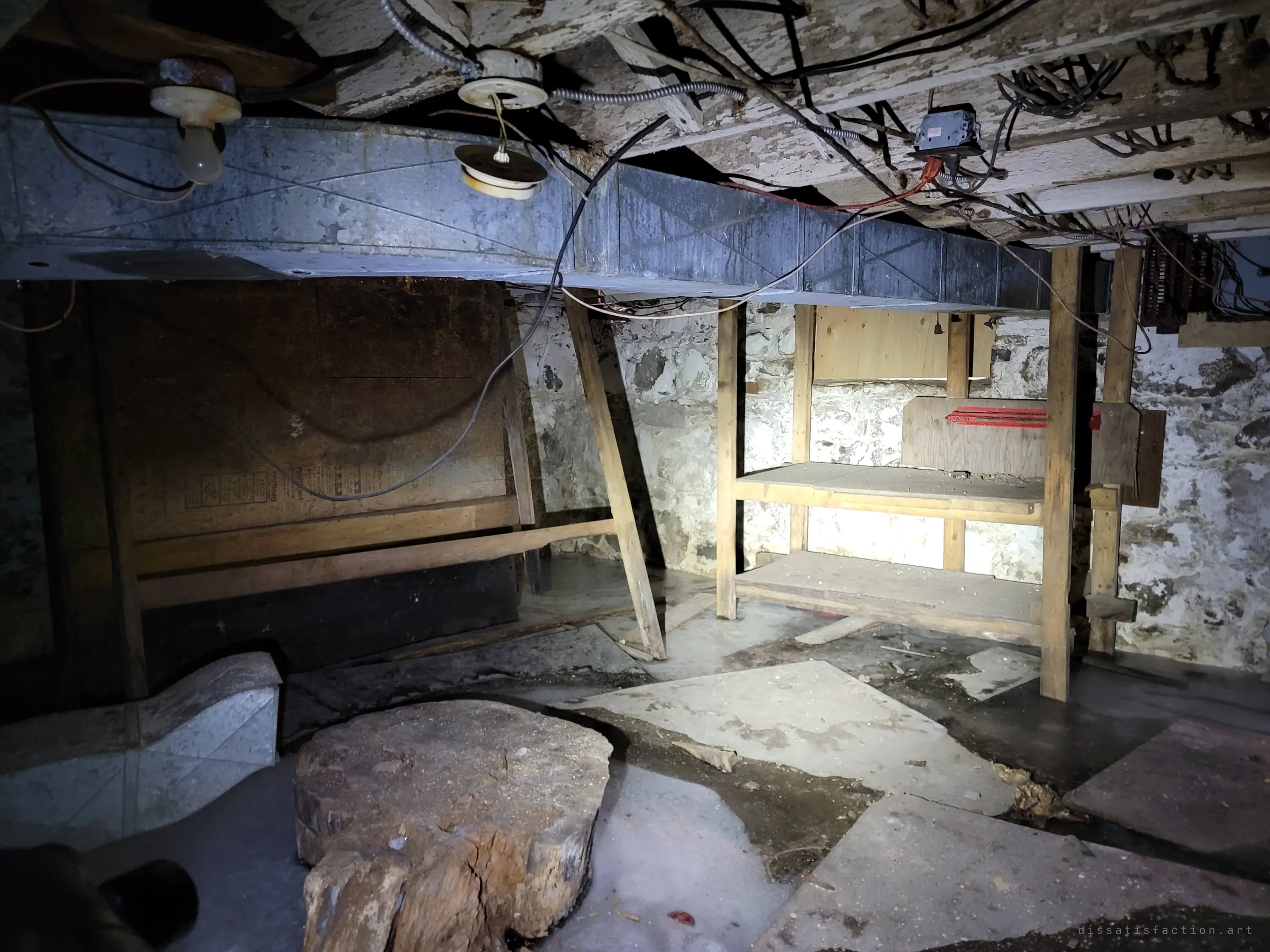 A flashlit basement with fieldstone walls, a sheet-metal duct and tangled wiring overhead, wooden shelving and an old stump on an icy floor. The rough stone foundation hints at the house's age.