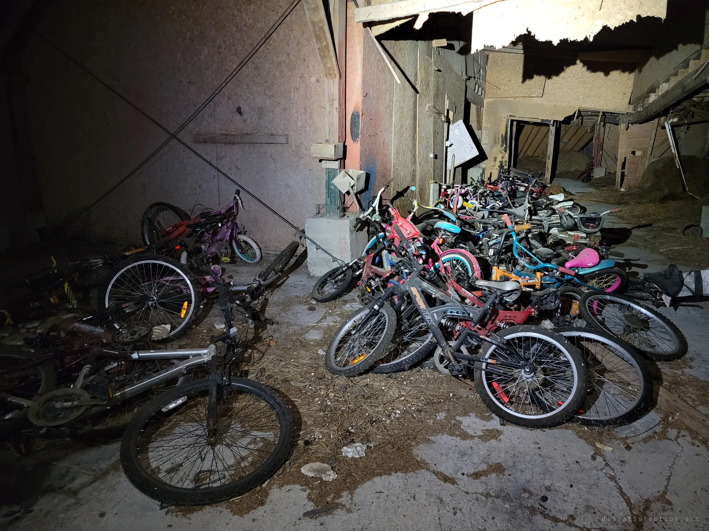 Dozens of abandoned children's and adult bicycles heaped against the walls of a dim barn aisle, torn board ceiling and hay in the stalls beyond. The sheer number of bikes is the story of the place.