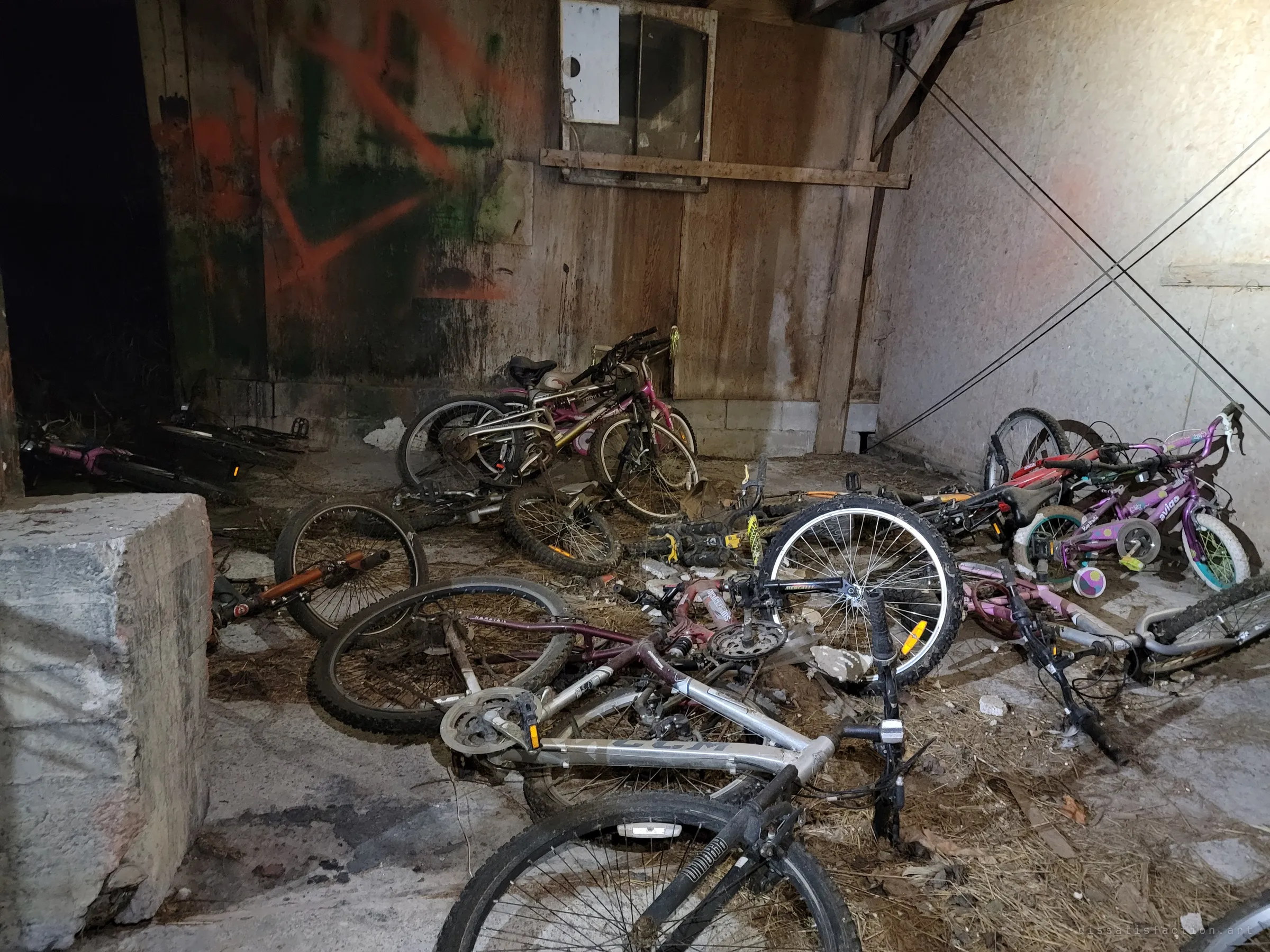 More bicycles tangled on a straw-strewn concrete floor beside a graffiti-sprayed plank wall and a boarded window. Kids' bikes with training wheels lie among worn mountain bikes.