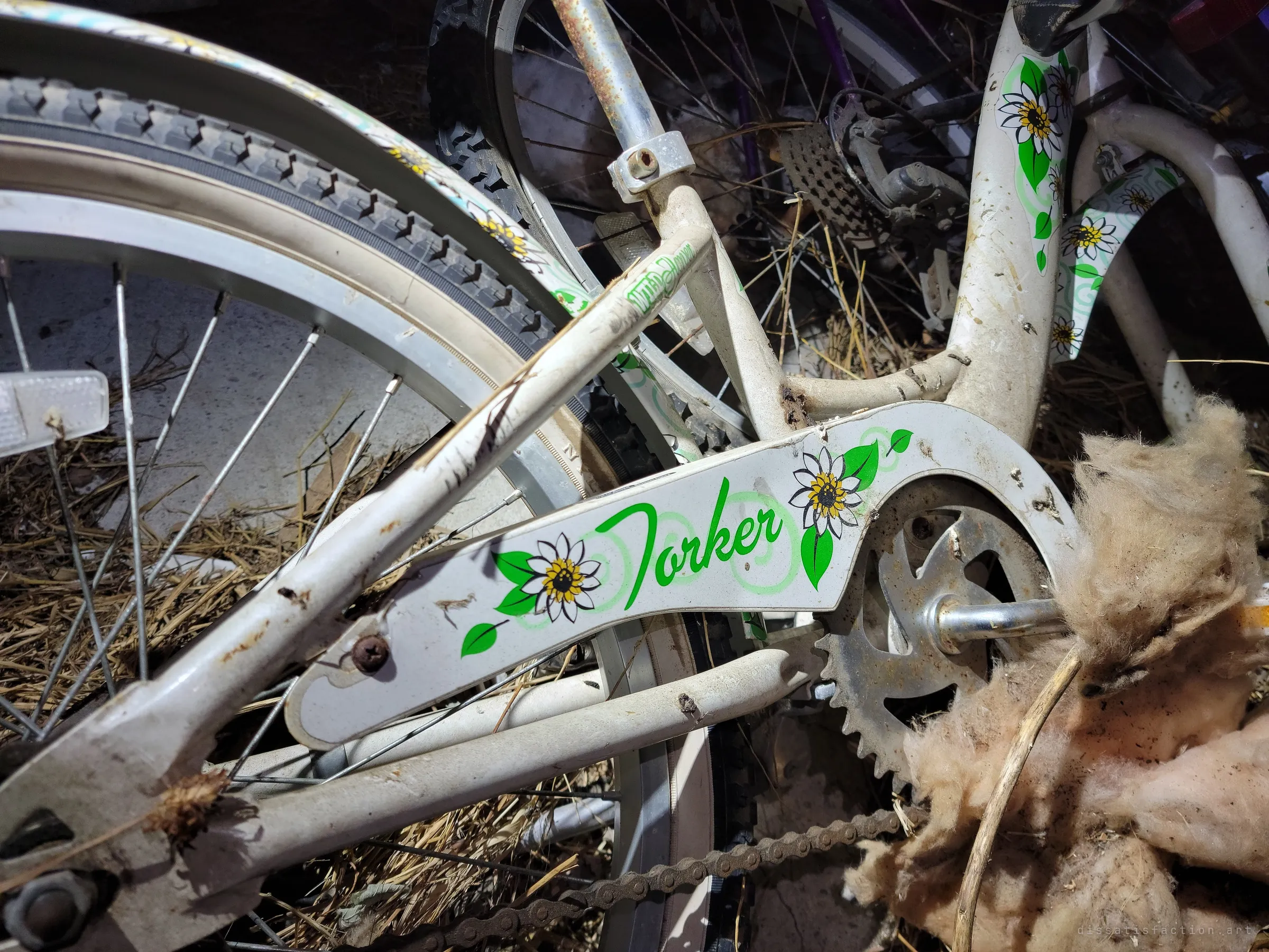 Close-up of a dusty white girl's bicycle with a daisy-decorated chain guard, lying in straw and pink insulation. A small, once-cheerful detail left to rot with the rest.