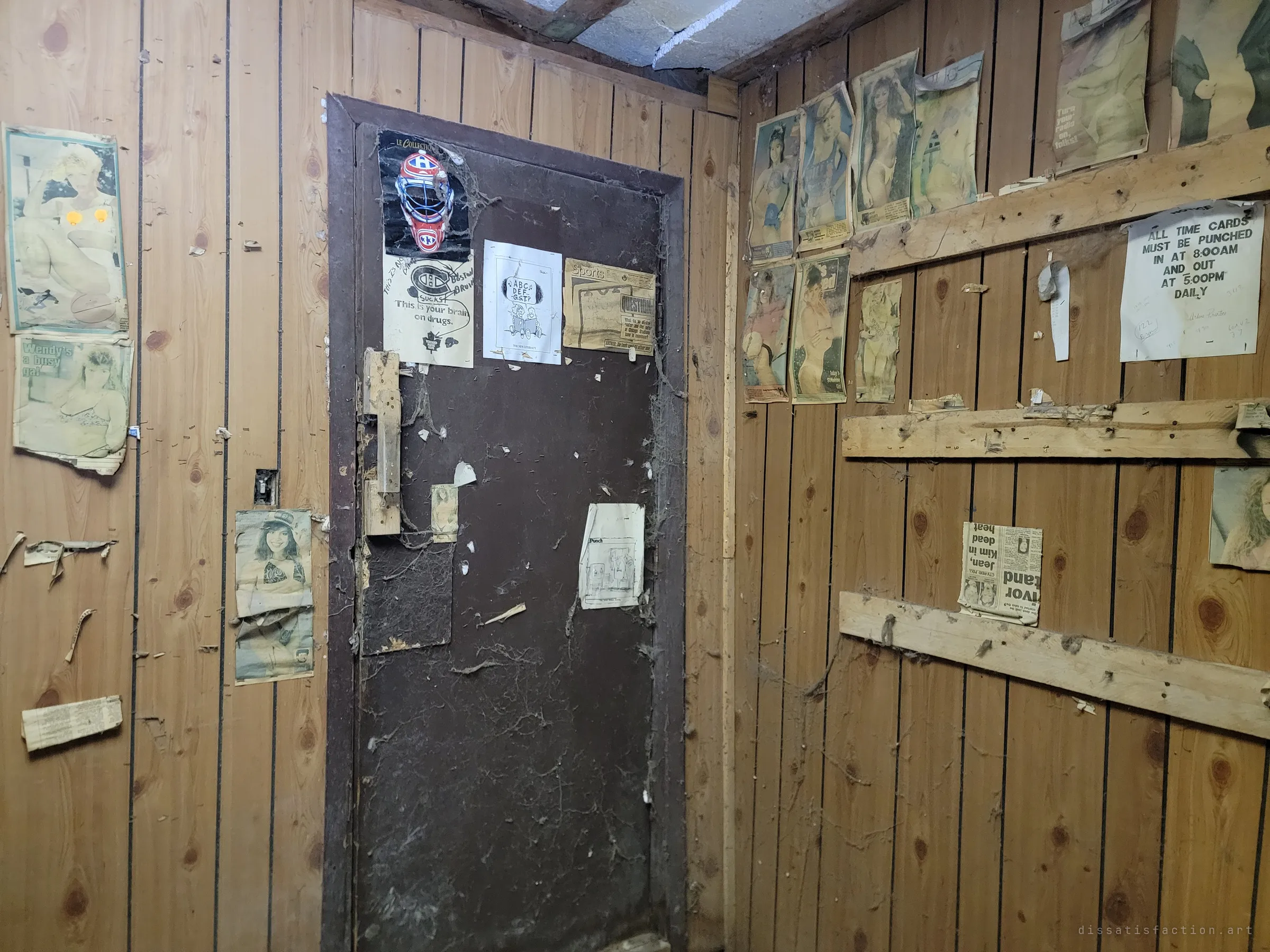 A wood-panelled break room with a dark door covered in cobwebs, old hockey clippings, cartoons and yellowed pin-up newspaper pages. A typed notice about punching time cards hints at the workers who once used it.