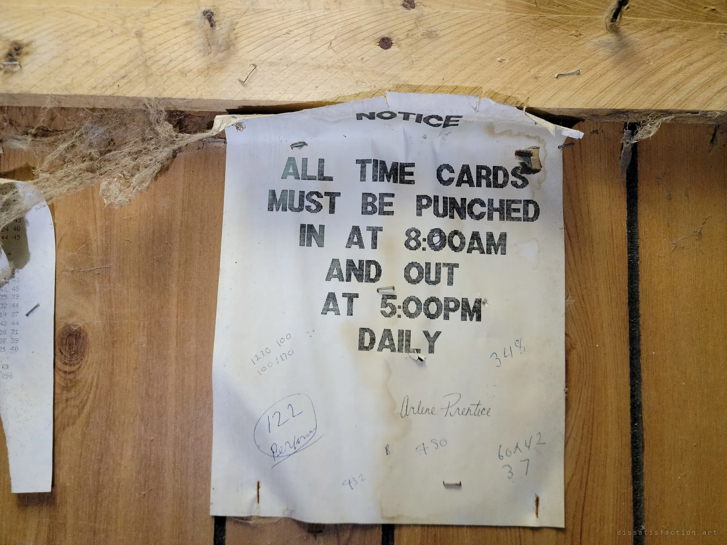 A stained typed notice stapled to the wall telling staff to punch their time cards in the morning and out in the afternoon, with pencilled numbers scrawled around it. A trace of the daily routine of a long-gone workplace.