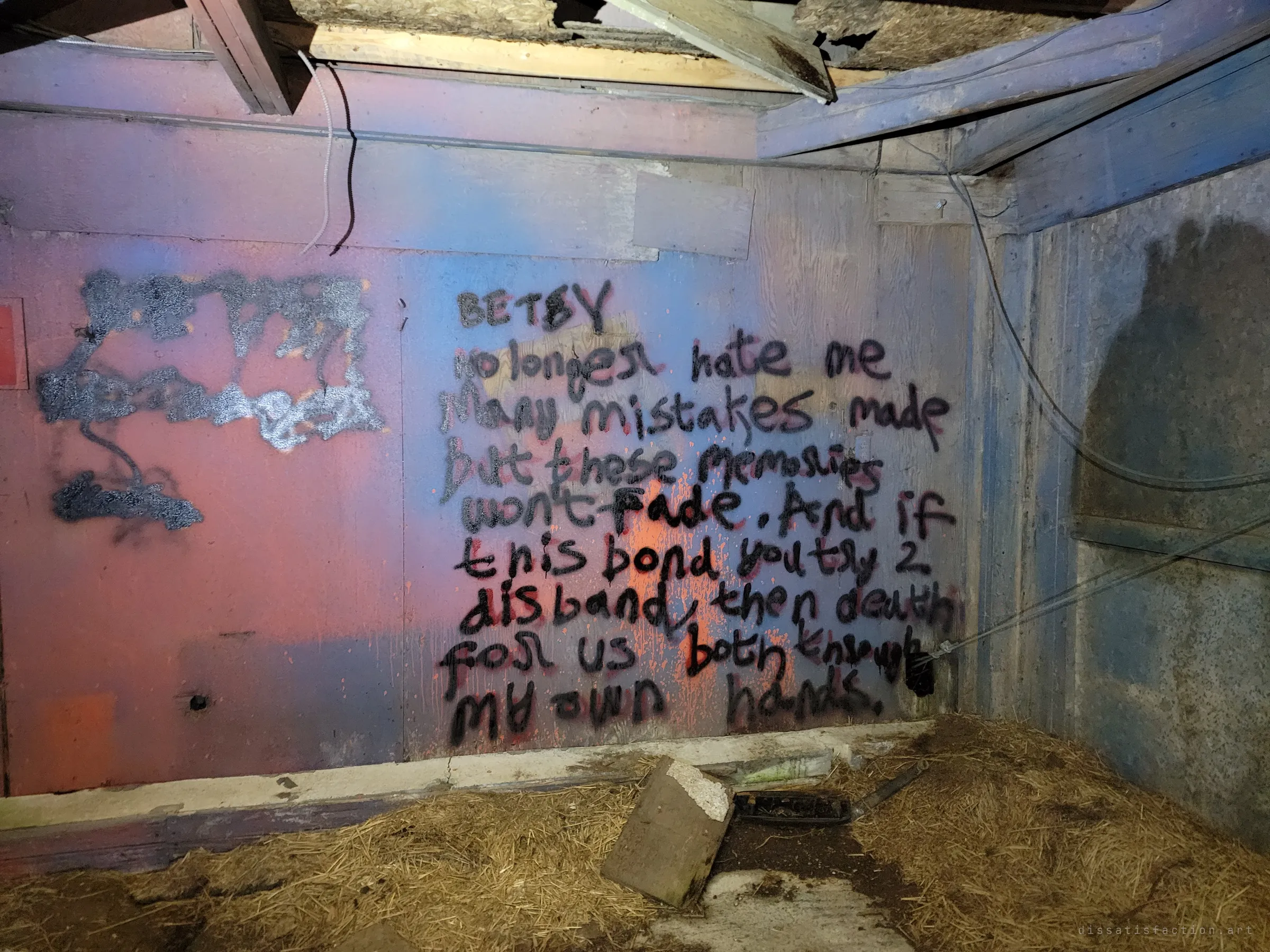A hand-sprayed graffiti poem about regret and a bond that must not break, lit pink and blue on a plywood wall above a straw-covered floor. Someone left a raw confession in the dark.