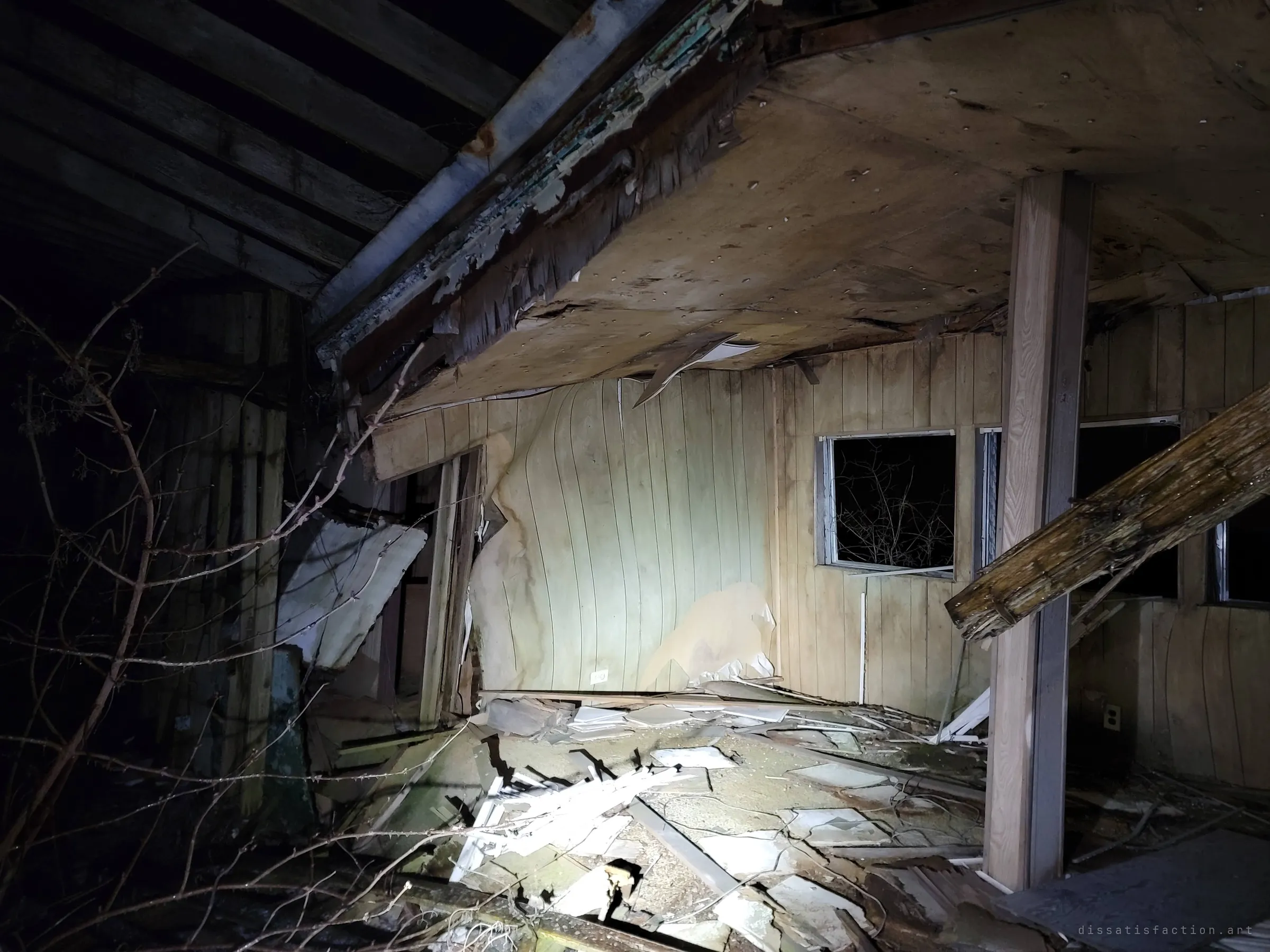 A collapsed ceiling sags into a panelled room, its floor buried in broken tiles and debris, with bare branches pushing in through the gaps. Nature is already moving back inside.