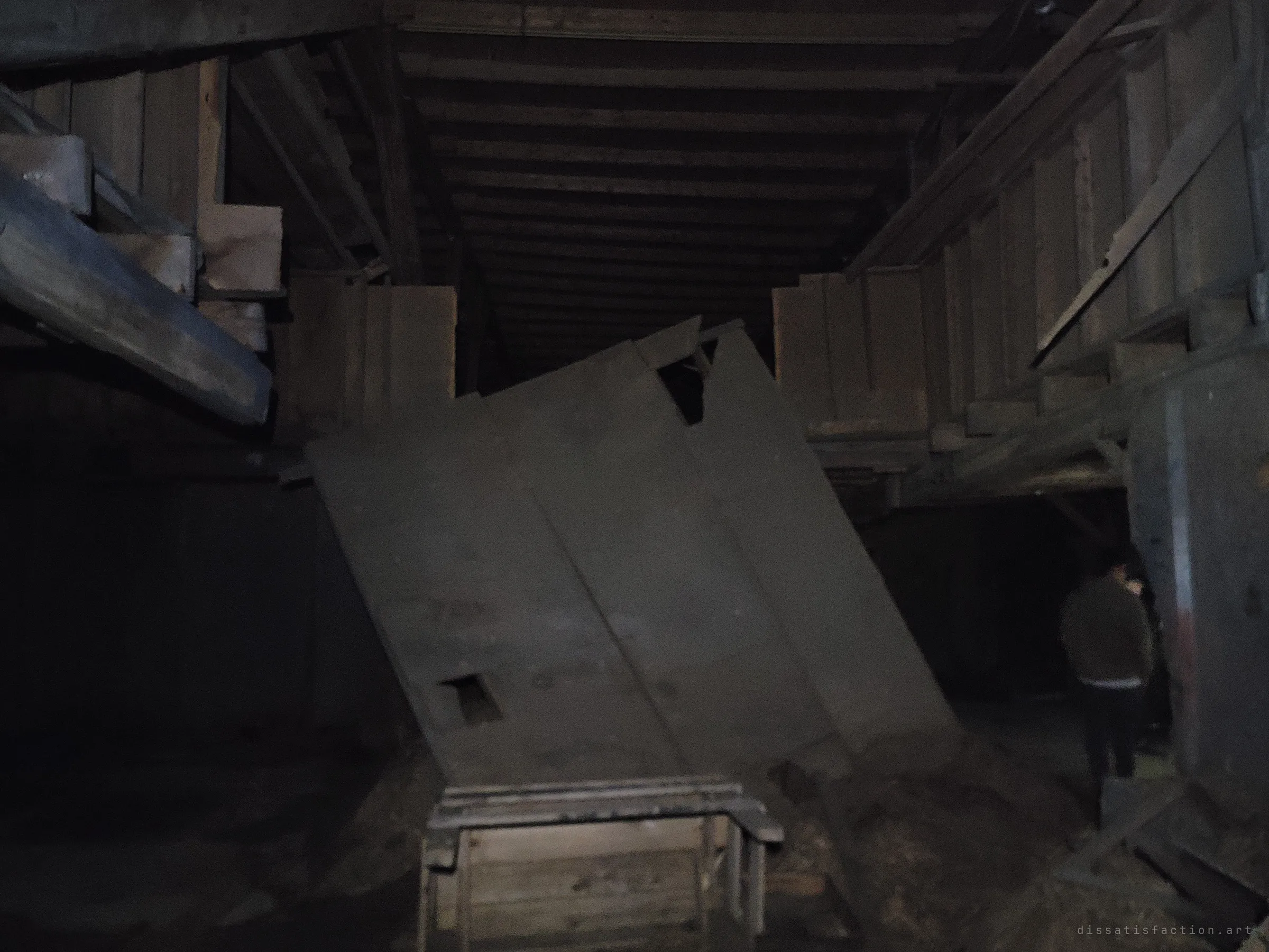 A long grey wooden chute or bin leans askew in a dim barn loft lined with plank bays, while a fellow explorer stands in the shadows at right. The scale of the timber structure shows in the gloom.