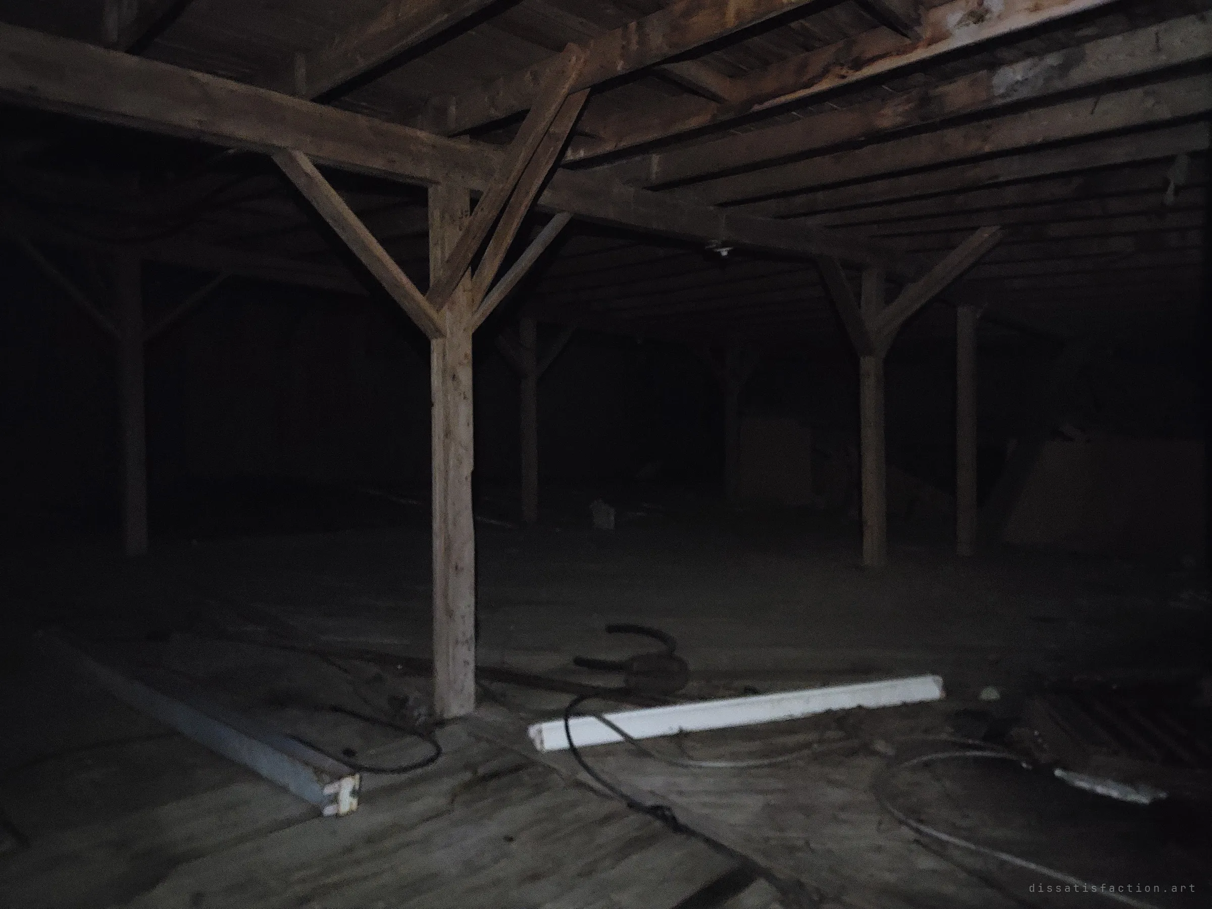Y-braced timber posts march into darkness across an empty barn floor strewn with cables and loose boards. The open span feels vast and silent.