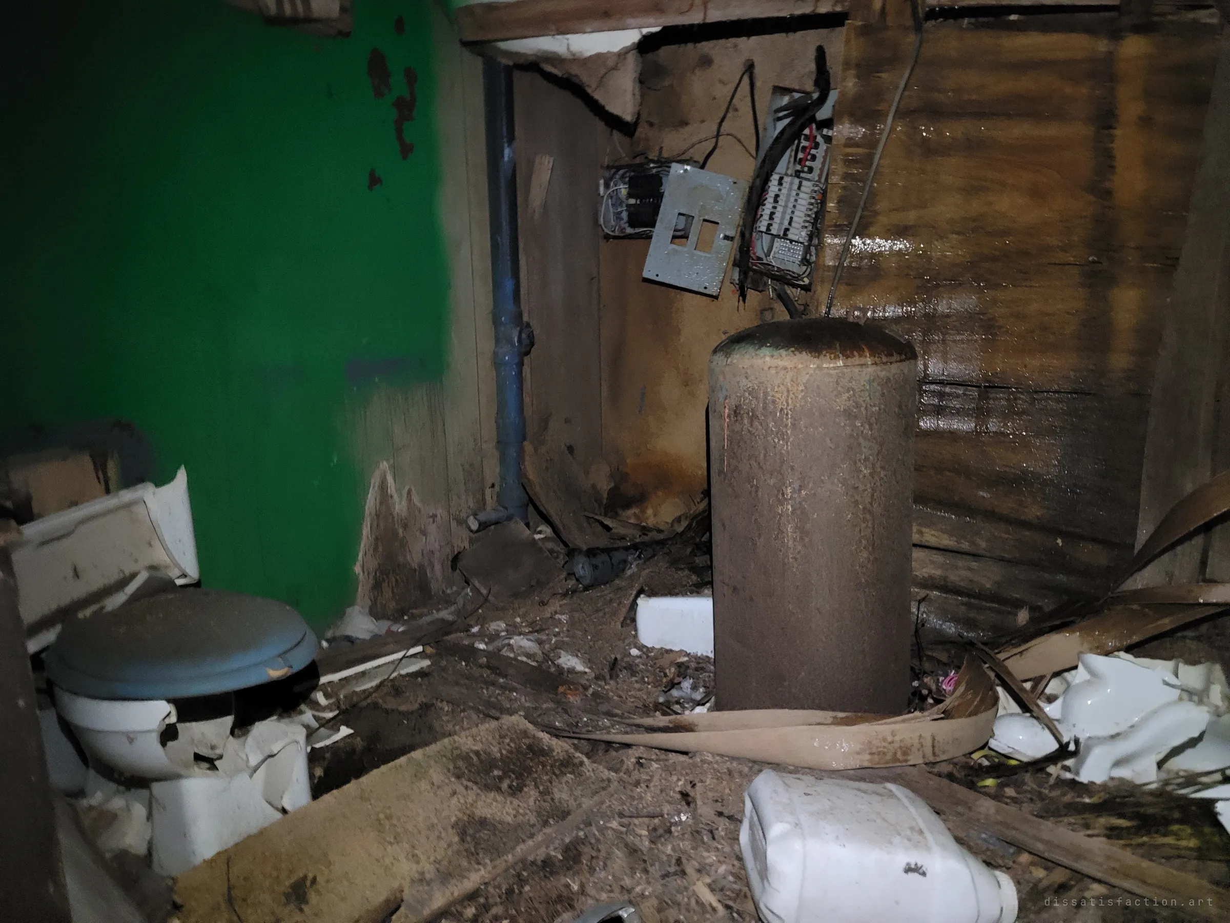 A rusted water tank stands among smashed porcelain beside a toilet and a gutted electrical panel against a bright green wall. The small washroom has been torn apart.