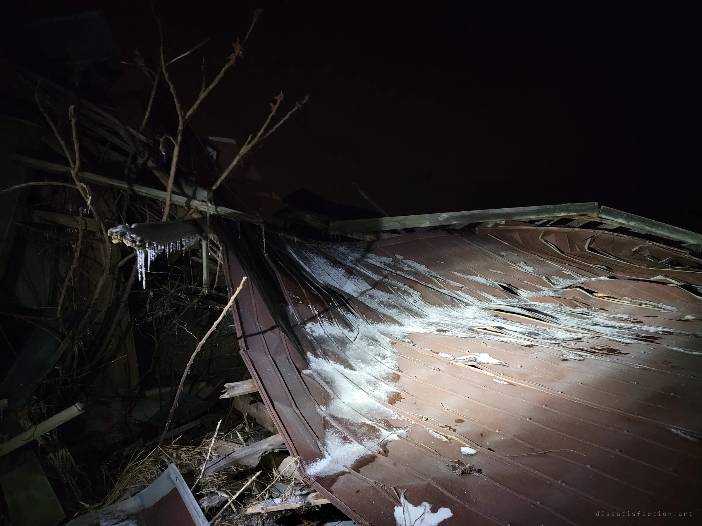 A crumpled red metal roof lies caved in under frost and icicles, tangled with bare branches against a black night sky. The building is folding in on itself.