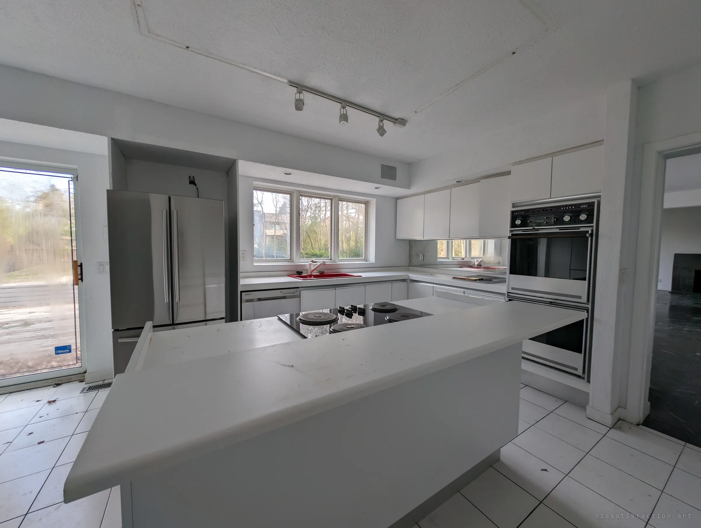 A bright all-white kitchen with an island cooktop, built-in double ovens and a red sink under the window. Everything is still in place, just dusty and abandoned.
