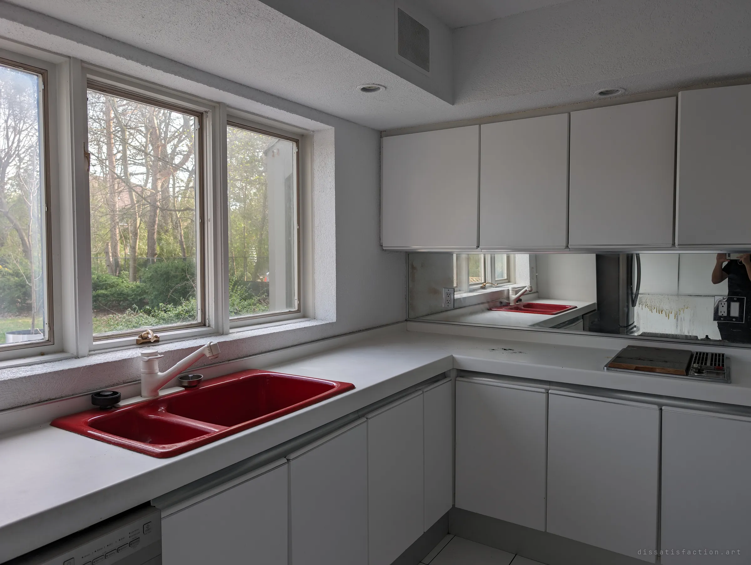 The red double sink beneath a row of windows, with the mirrored backsplash catching the photographer mid-shot. A small splash of colour in an otherwise white kitchen.