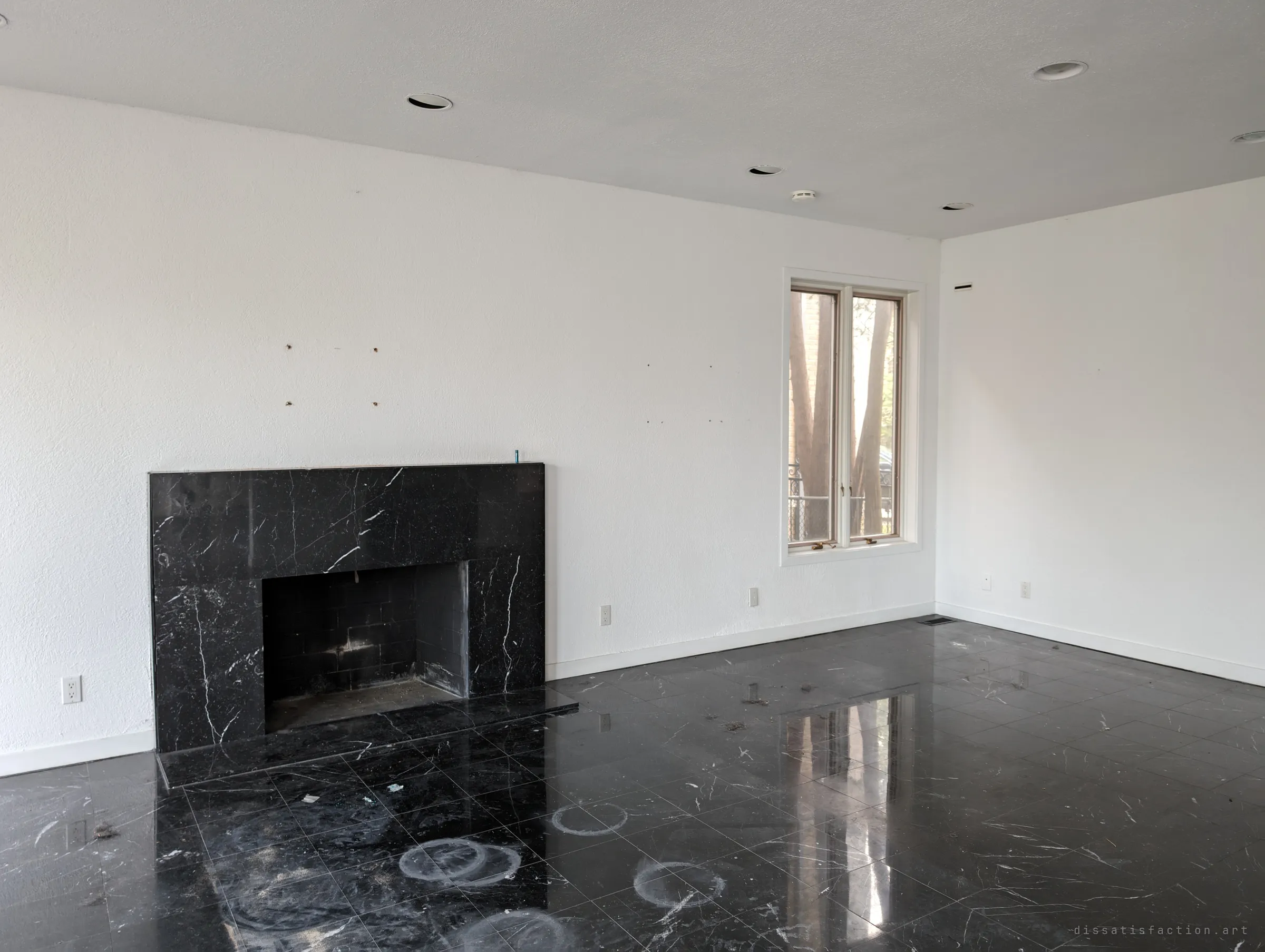 A black marble fireplace against a bare white wall, with pale rings left on the glossy floor where furniture or pots once stood. Screw holes mark where something used to hang.