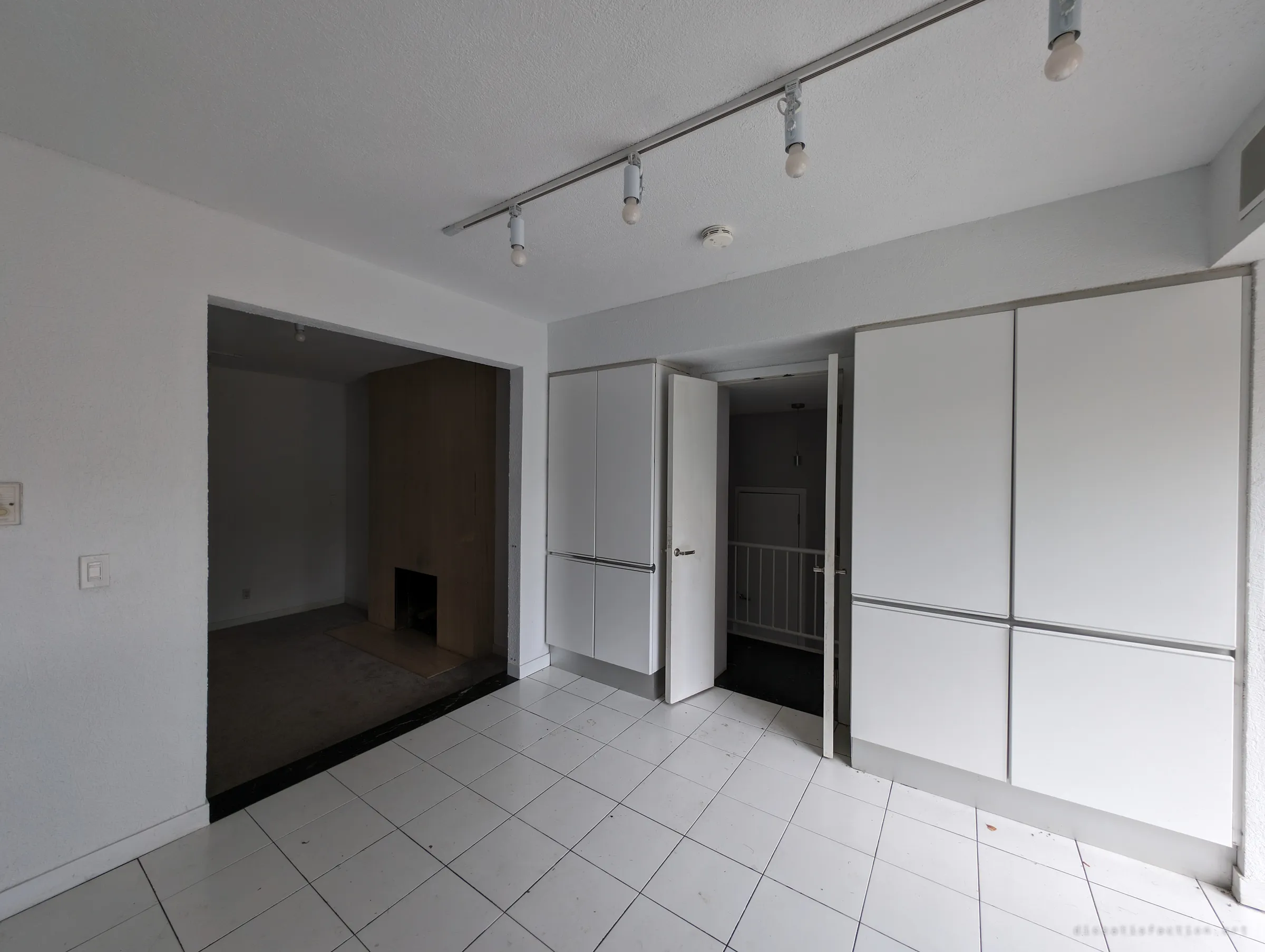 A white-tiled room lined with tall flush cabinets, a door left ajar onto a stair landing and a dark sunken den beyond. Track lights with bare bulbs still hang from the ceiling.