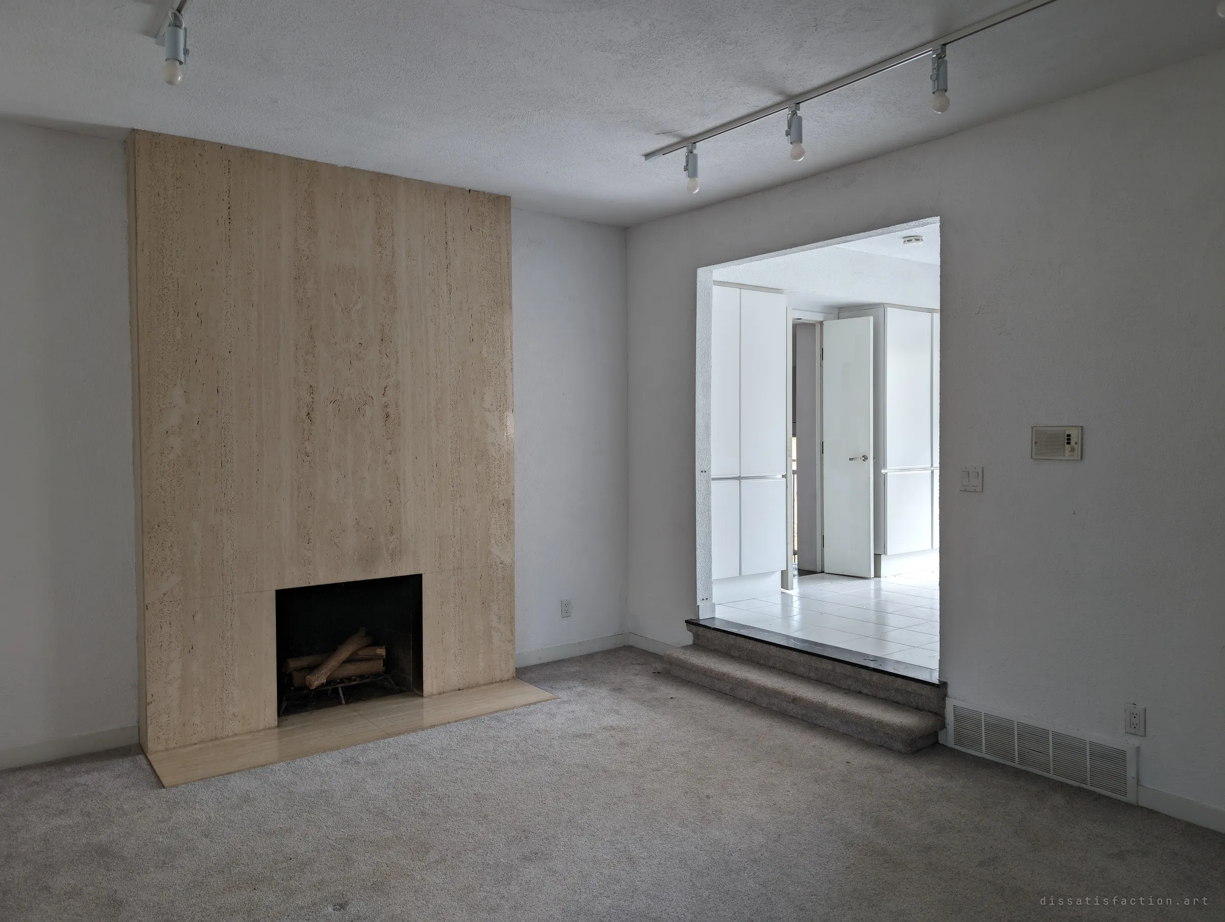 The carpeted den with a tall travertine fireplace, logs still waiting in the grate, looking back up the steps into the white room. The house feels paused rather than ruined.
