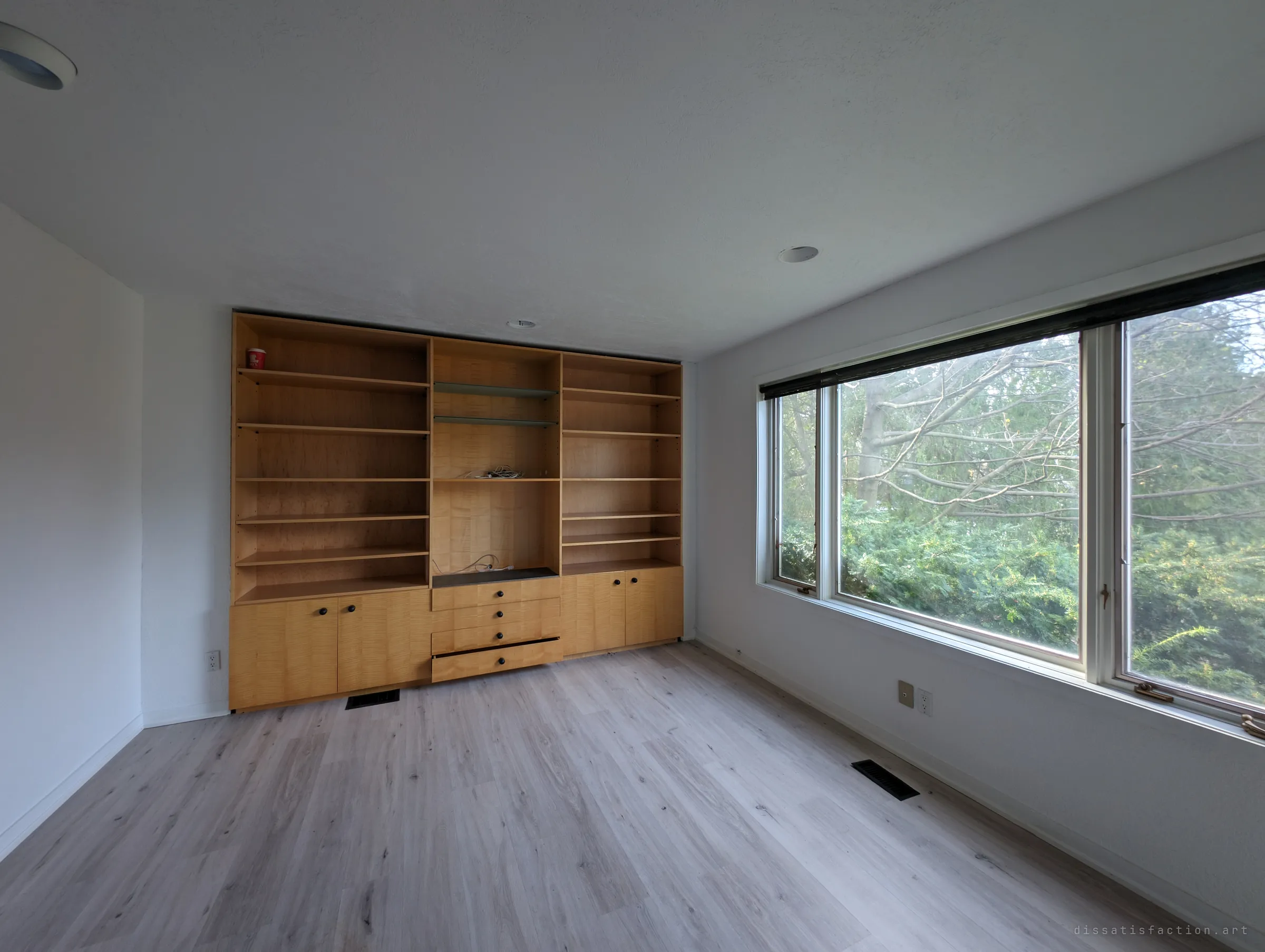 An empty room with a wall of built-in maple shelving and a wide window full of green branches. A single cup left on a top shelf is all that remains.