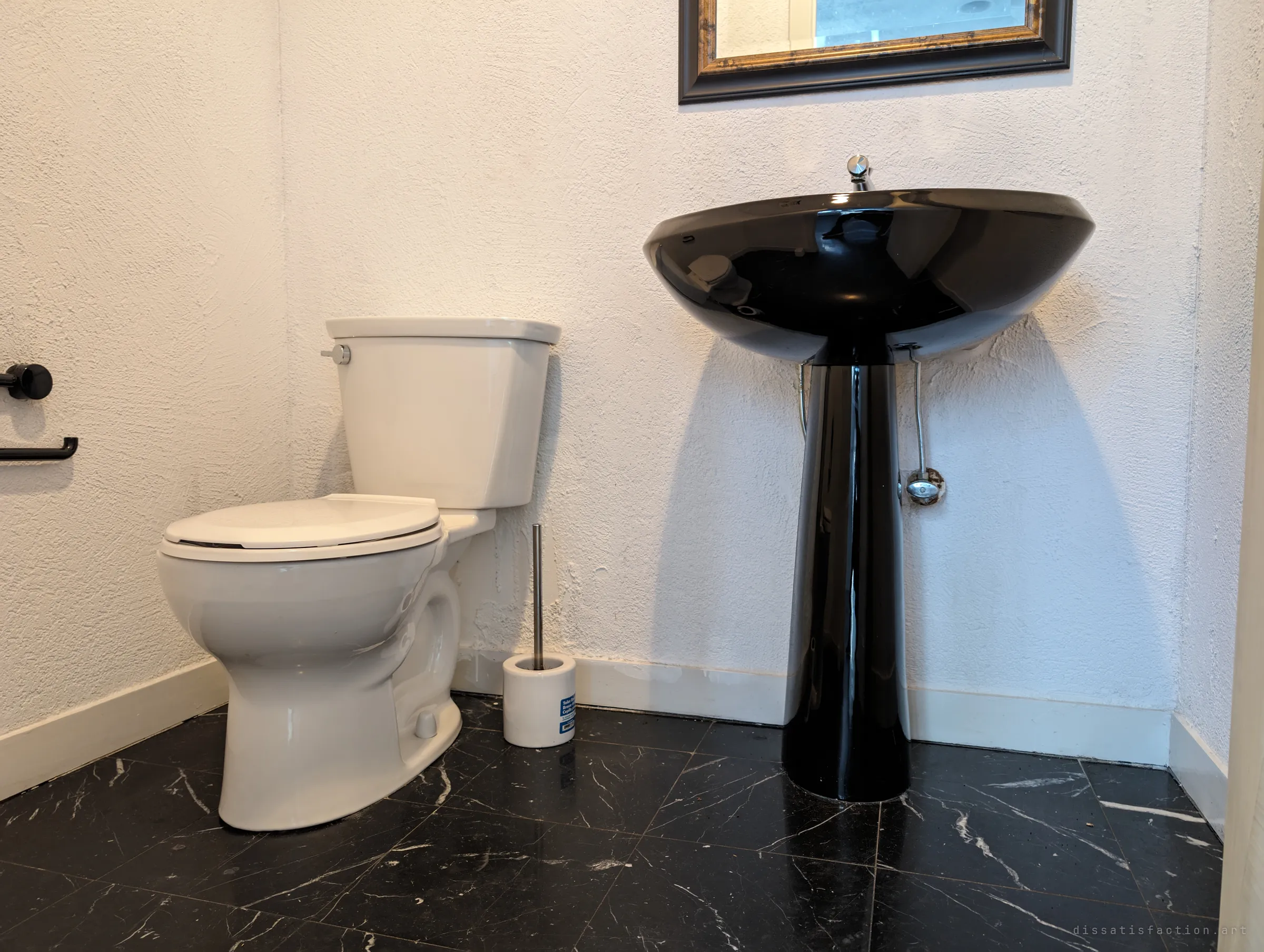 A powder room with a glossy black pedestal sink and a white toilet on black marble-look tile. The fixtures are still in place and oddly clean, as if the house was only just left.