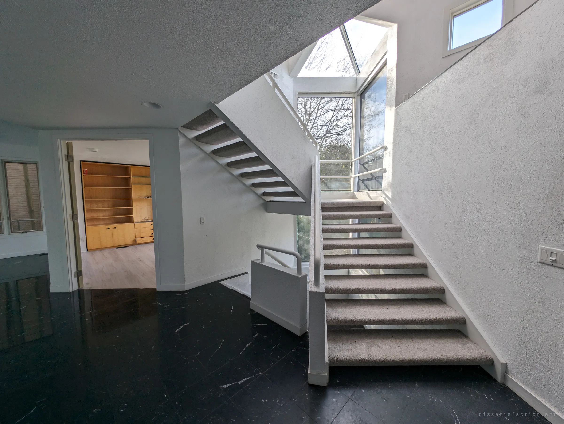 A white open-riser staircase with grey carpeted treads climbs toward a glass-roofed stairwell full of bare trees. Through a doorway, empty built-in wood shelving waits in a bare study.