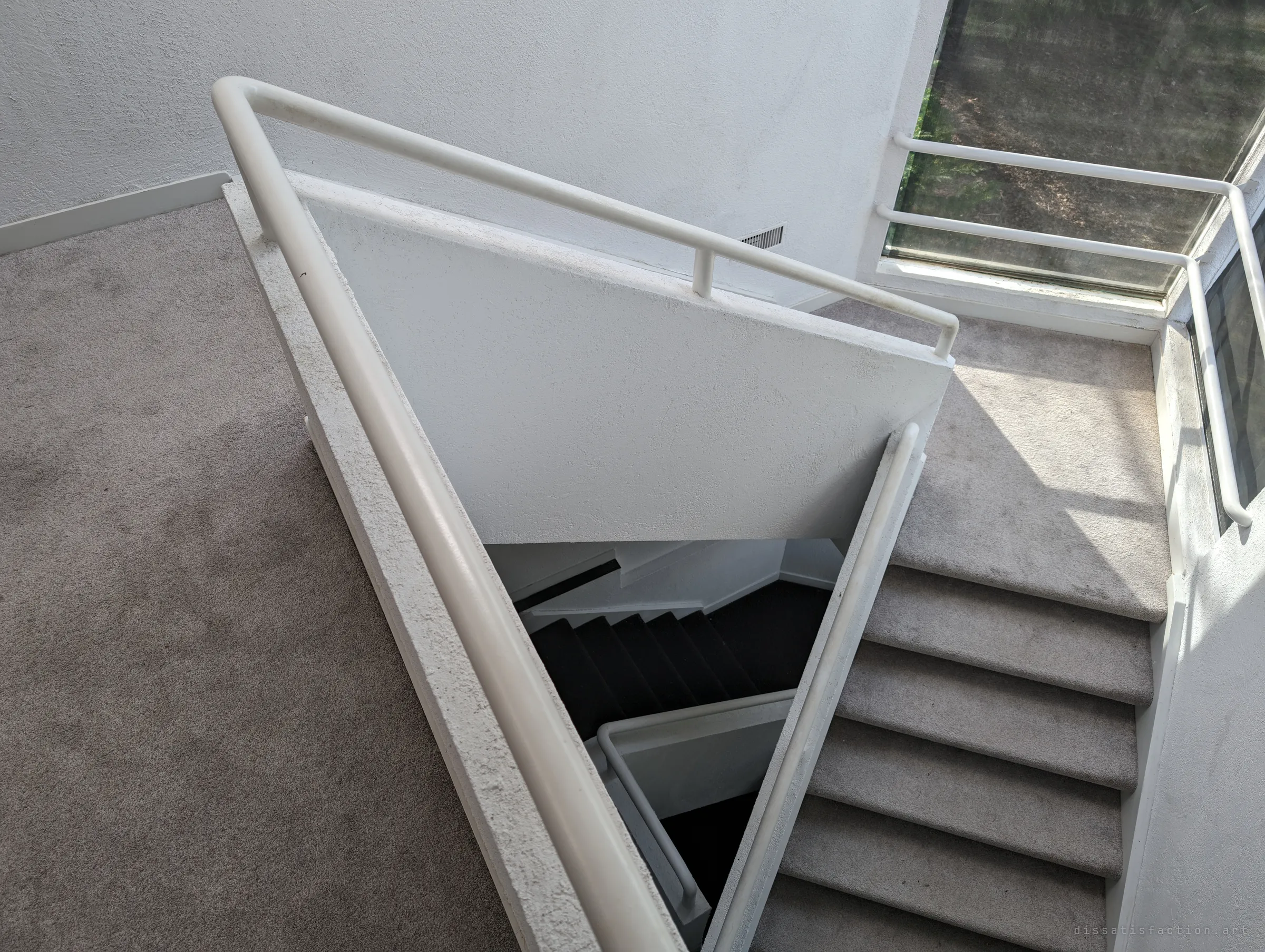 Looking down the stairwell from the upper landing, white tube railings frame a triangular drop into the dark lower flights. The geometry shows how the house was designed around this central void.