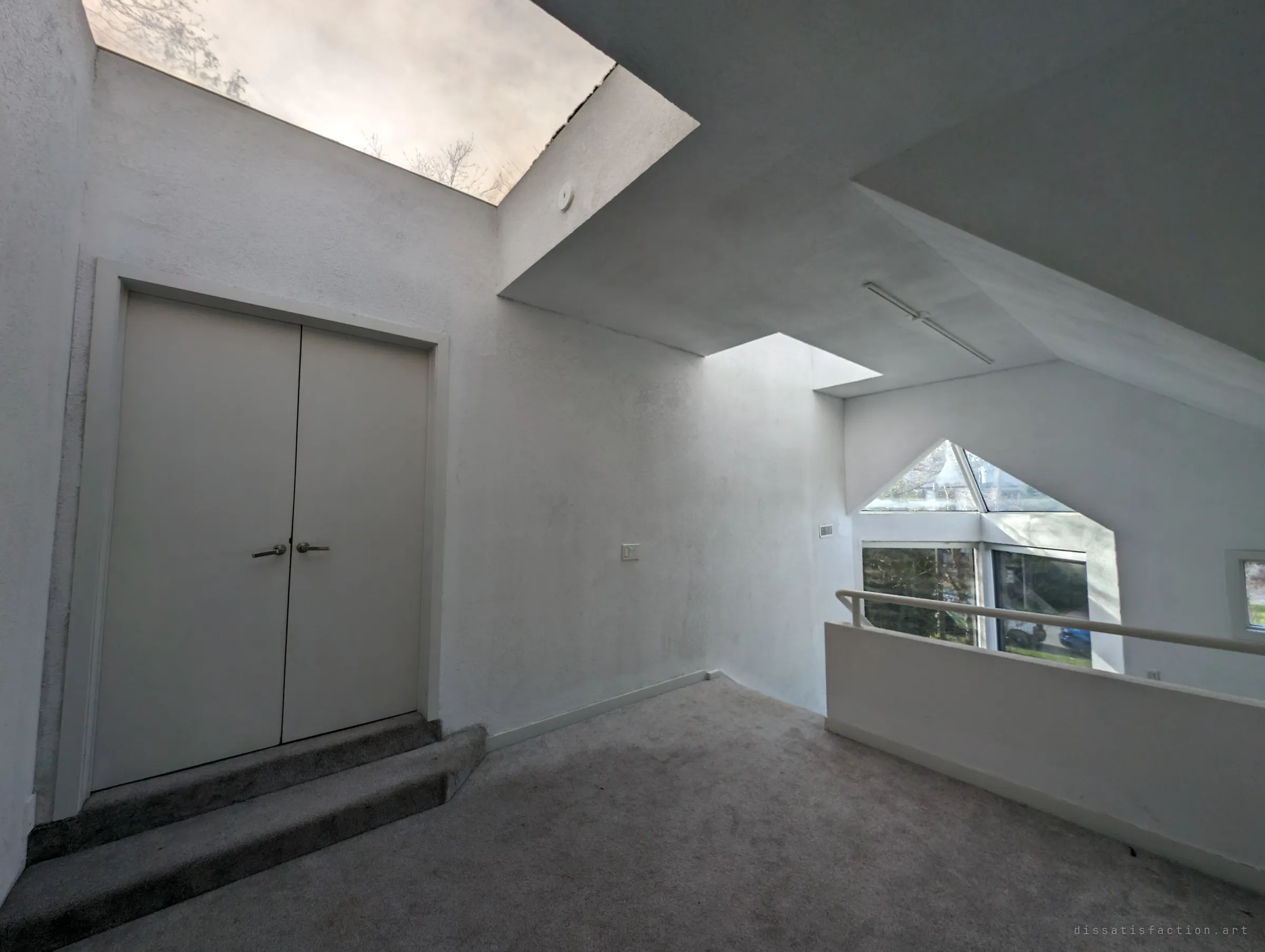 An upper landing with white double doors under a cut-out skylight, and a pointed glass bay window beyond the railing. The angular, all-white interior gives the house its gallery-like feel.