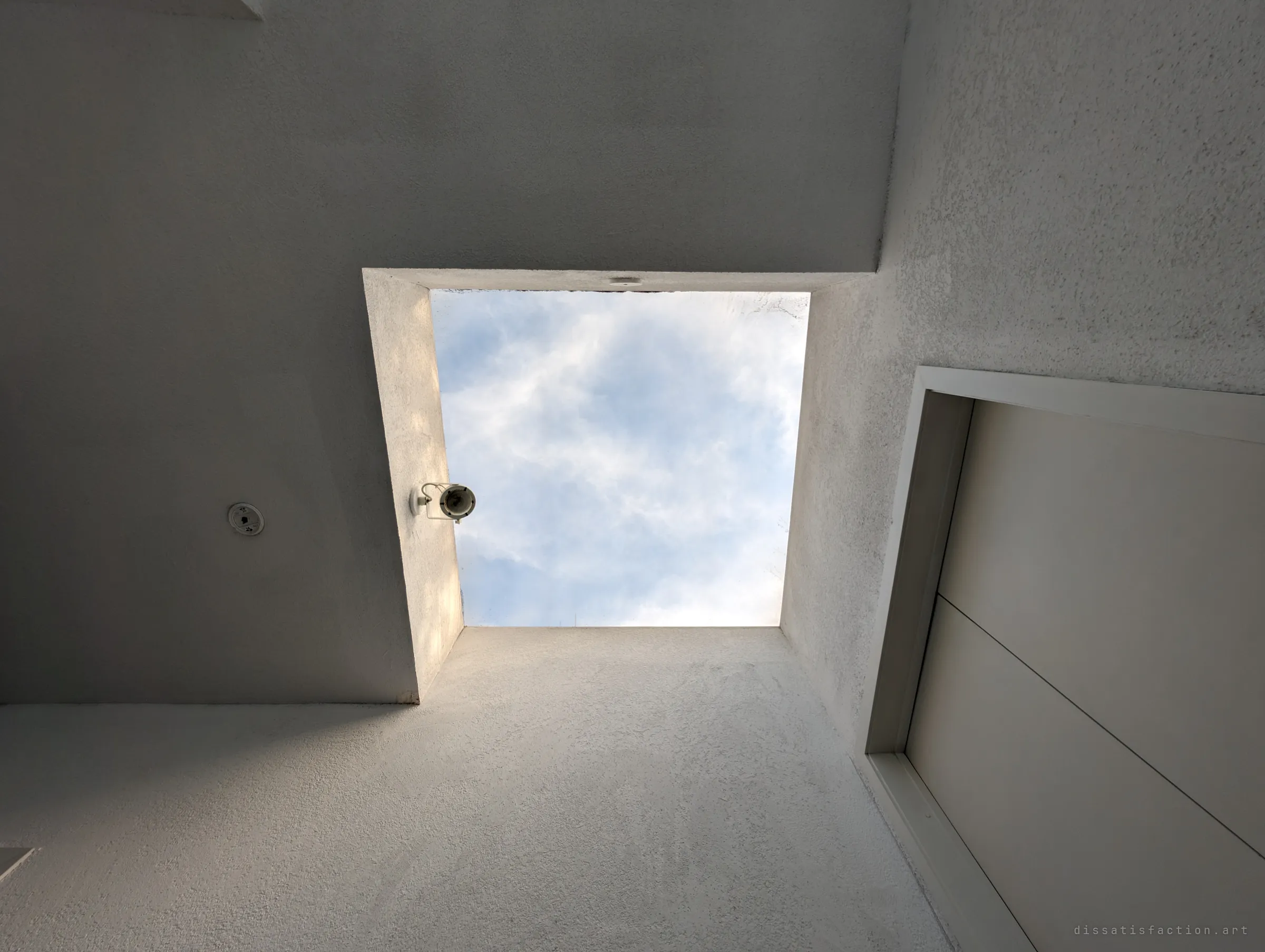 Looking straight up into a square skylight shaft at a pale cloudy sky, with a small spotlight clinging to one edge. A quiet detail of the light-filled design.