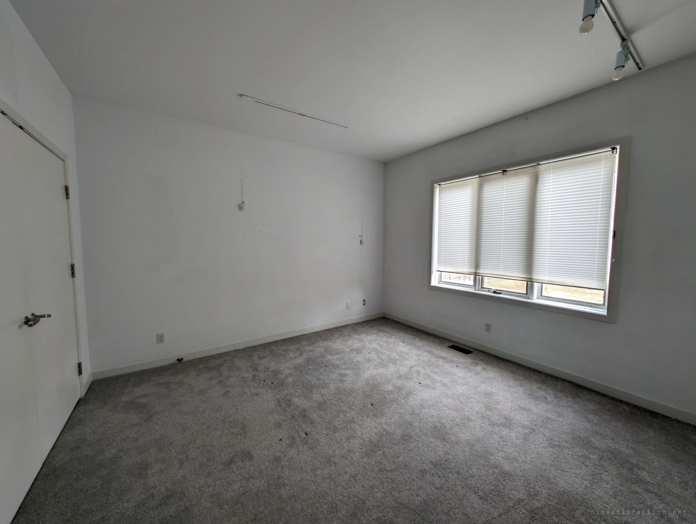 An empty carpeted bedroom with pleated blinds drawn over a wide window and bare track lighting on the ceiling. Faint marks on the wall show where things once hung.