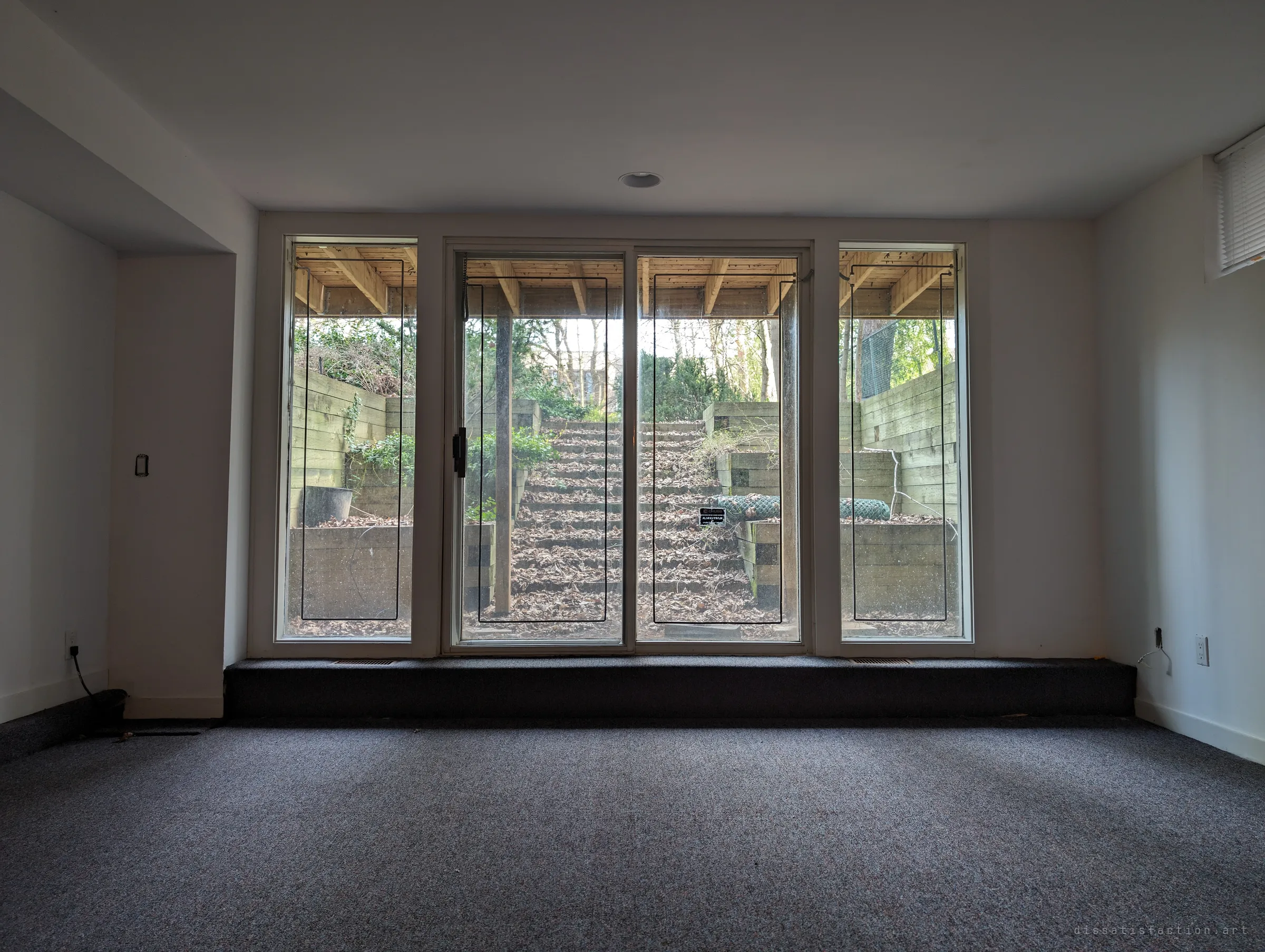 A walk-out basement room looks through tall windows onto timber retaining walls and leaf-covered steps climbing out of a sunken yard. The ground floor deck roofs over the view.