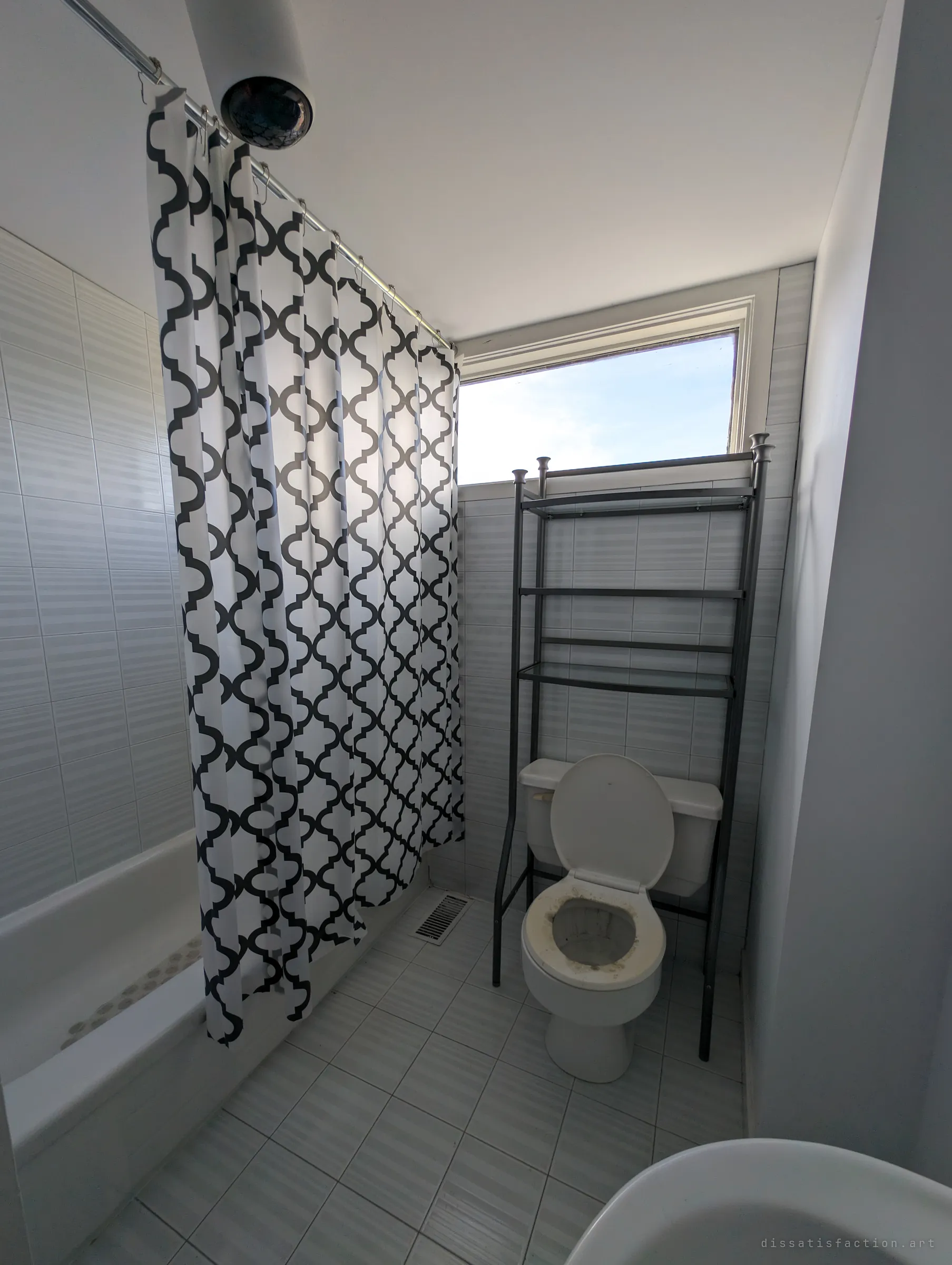 A small white-tiled bathroom with a patterned shower curtain, an over-toilet shelf and a high window showing only sky. The clean, staged fixtures sit oddly against the dust in the toilet bowl.