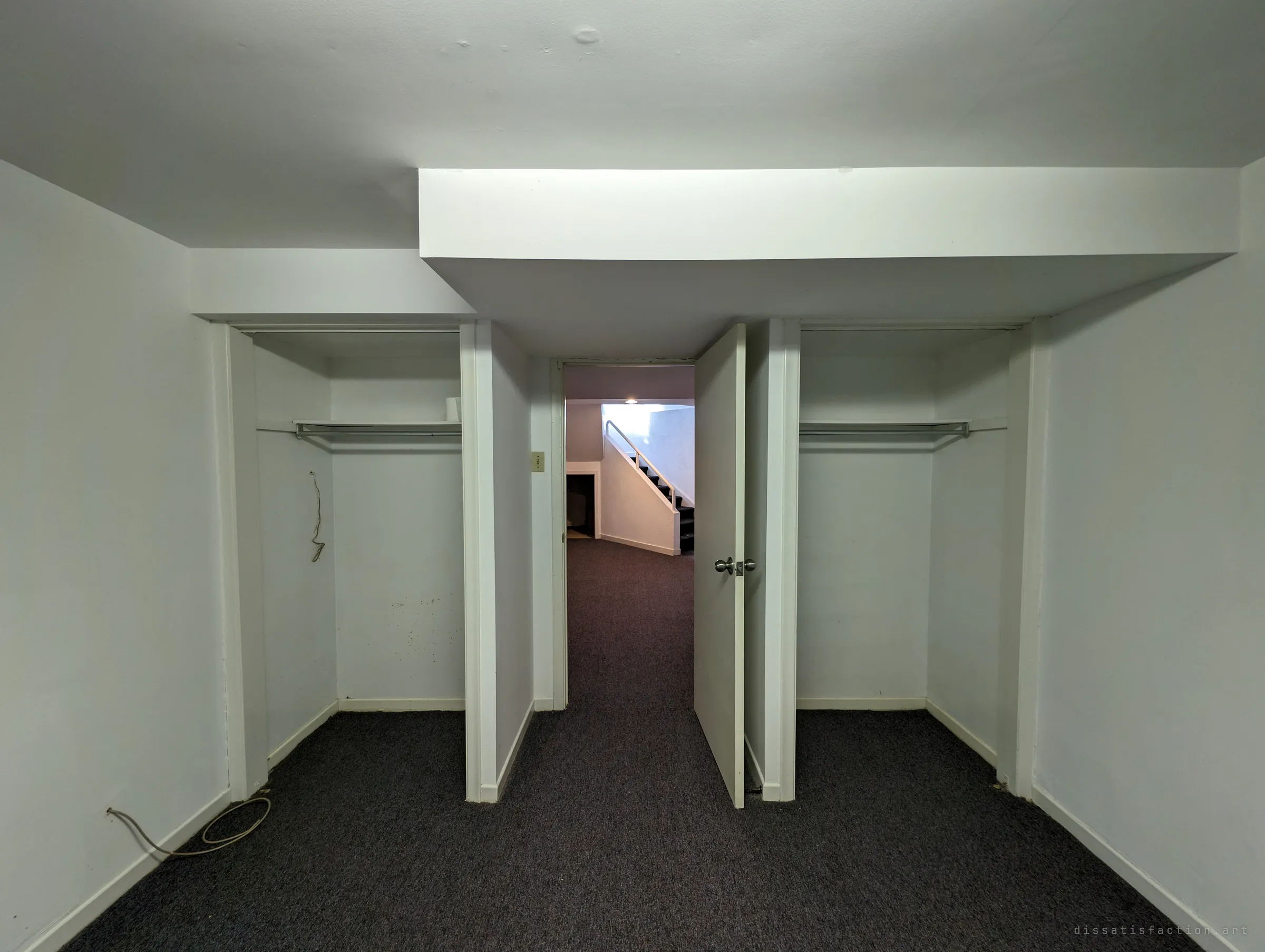 A carpeted basement bedroom with two empty open closets flanking a doorway that looks out to a stairwell. The symmetry and bare rails make the house feel recently emptied.