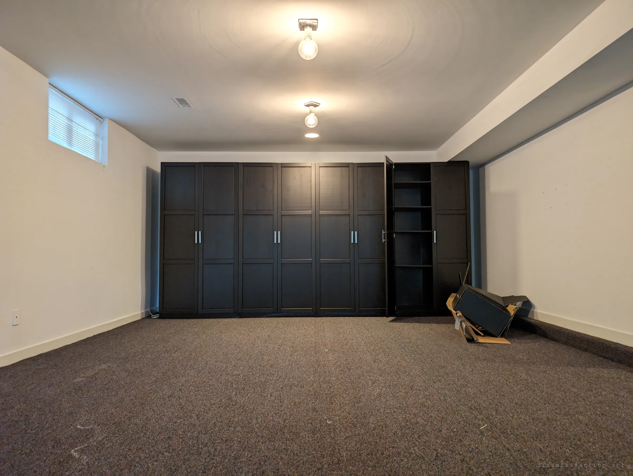 A long basement room lit by two bare bulbs, a wall of dark wardrobes with one door hanging open and a toppled cabinet in its box. Everything useful is gone, only the heavy furniture stayed.
