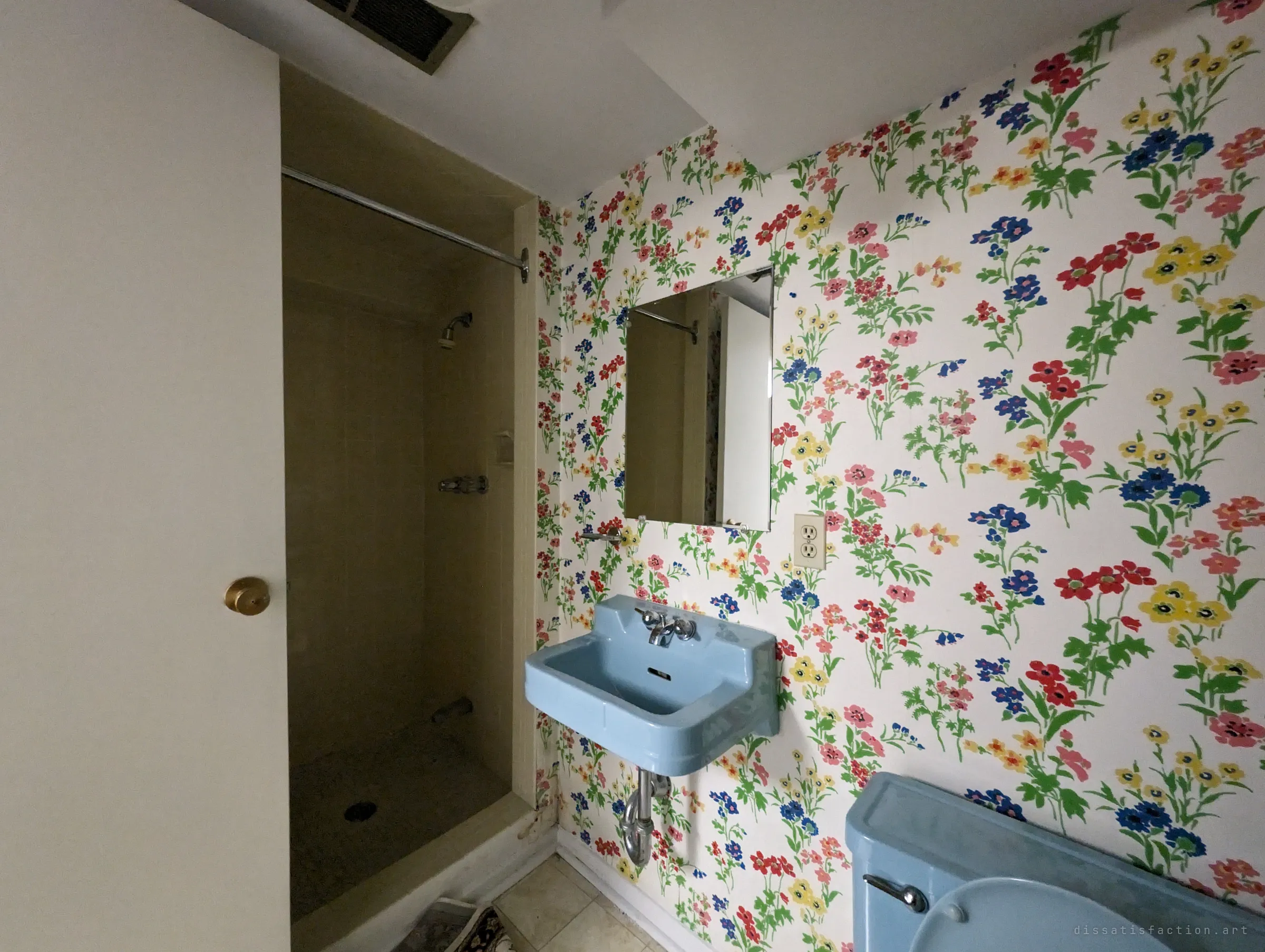 A bathroom papered in bright wildflowers, with a powder-blue wall sink and toilet beside a dim tiled shower stall. A vivid mid-century room left untouched by later renovations.