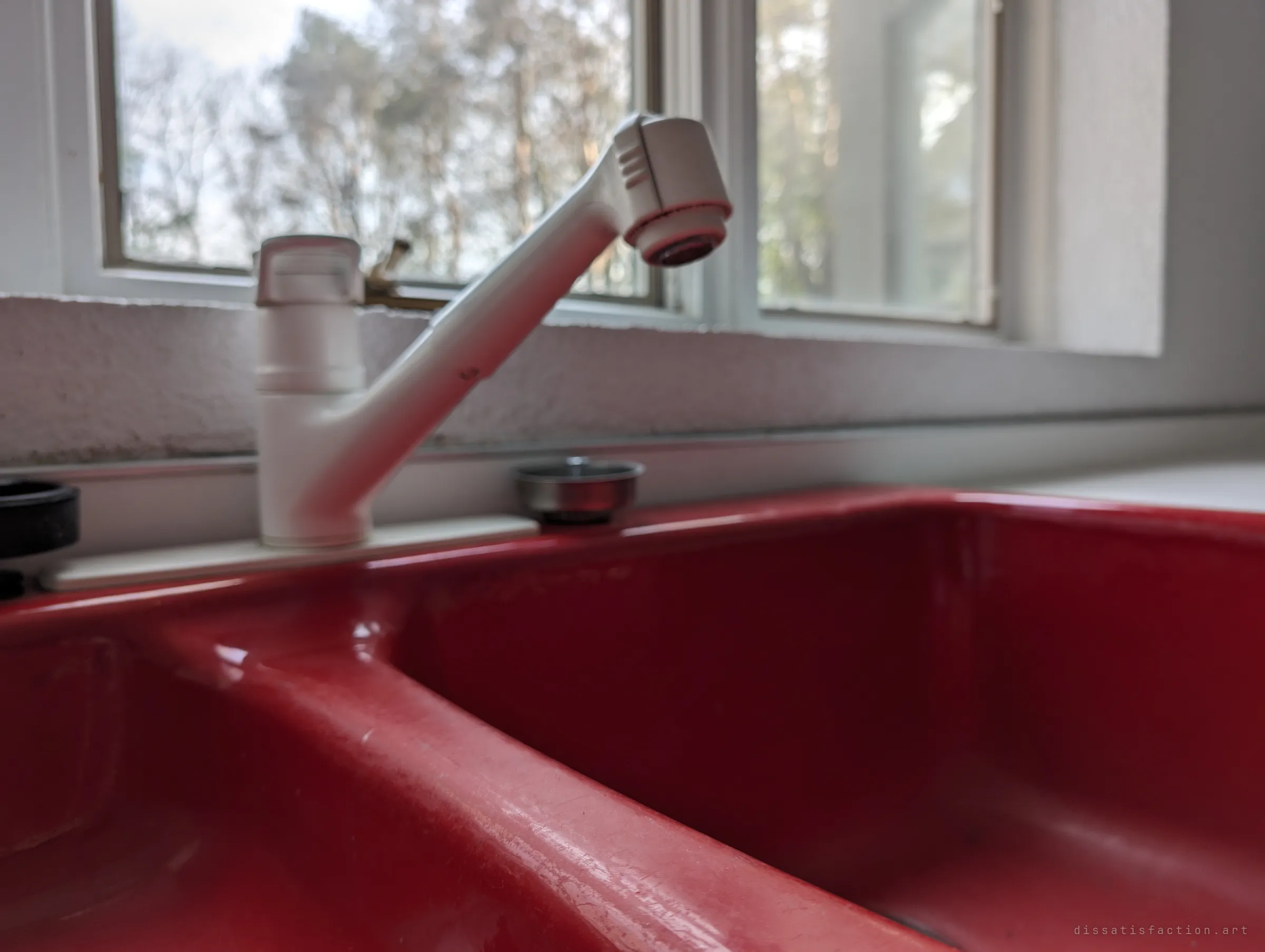 A glossy red double kitchen sink with a white pull-out faucet, bare trees soft in the window behind. A bold splash of colour in an otherwise faded kitchen.