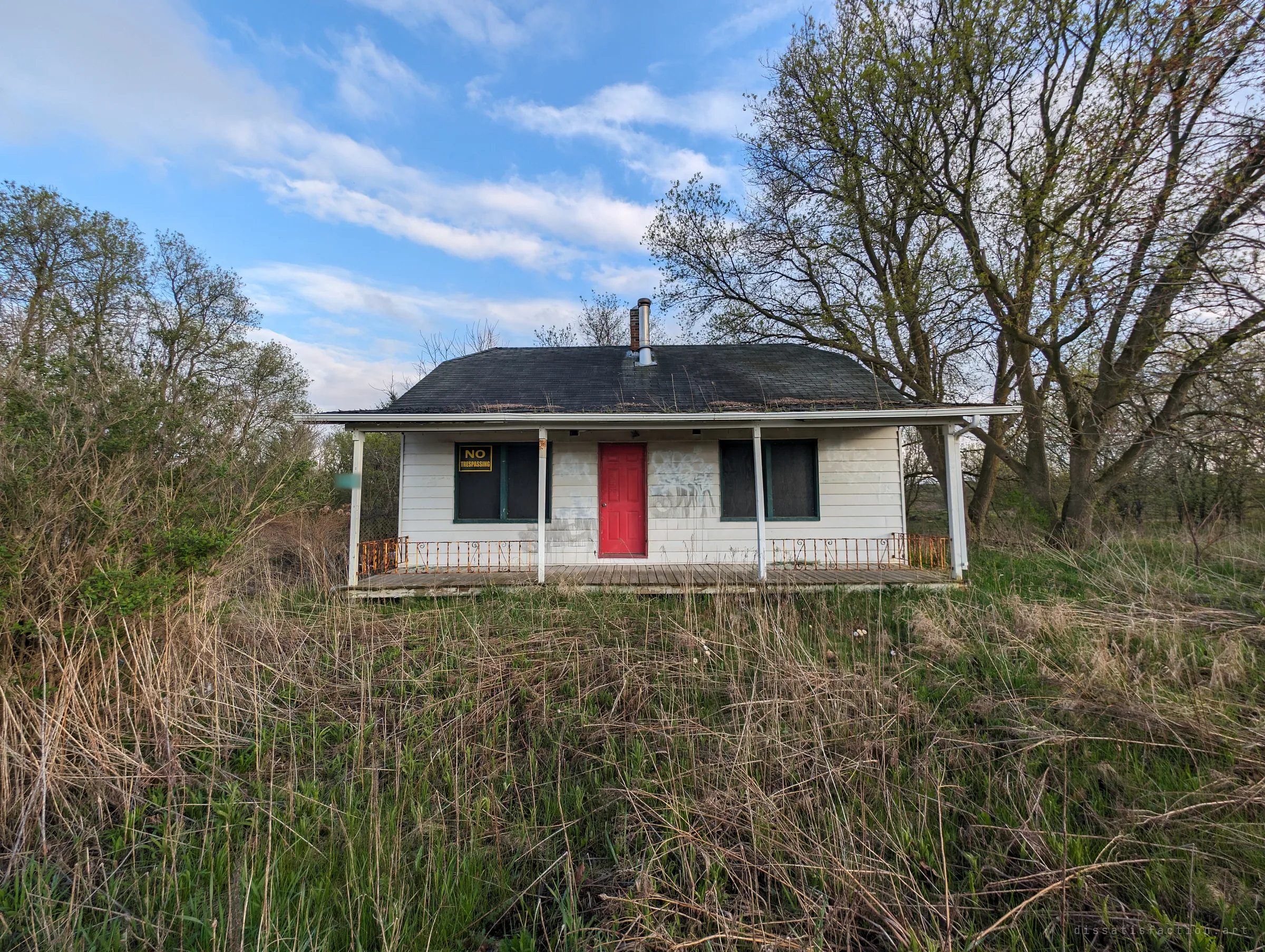 A small white single-storey house with a bright red door and a no-trespassing sign sits behind overgrown grass under a blue sky. It sets the scene: a modest home left to the weeds.