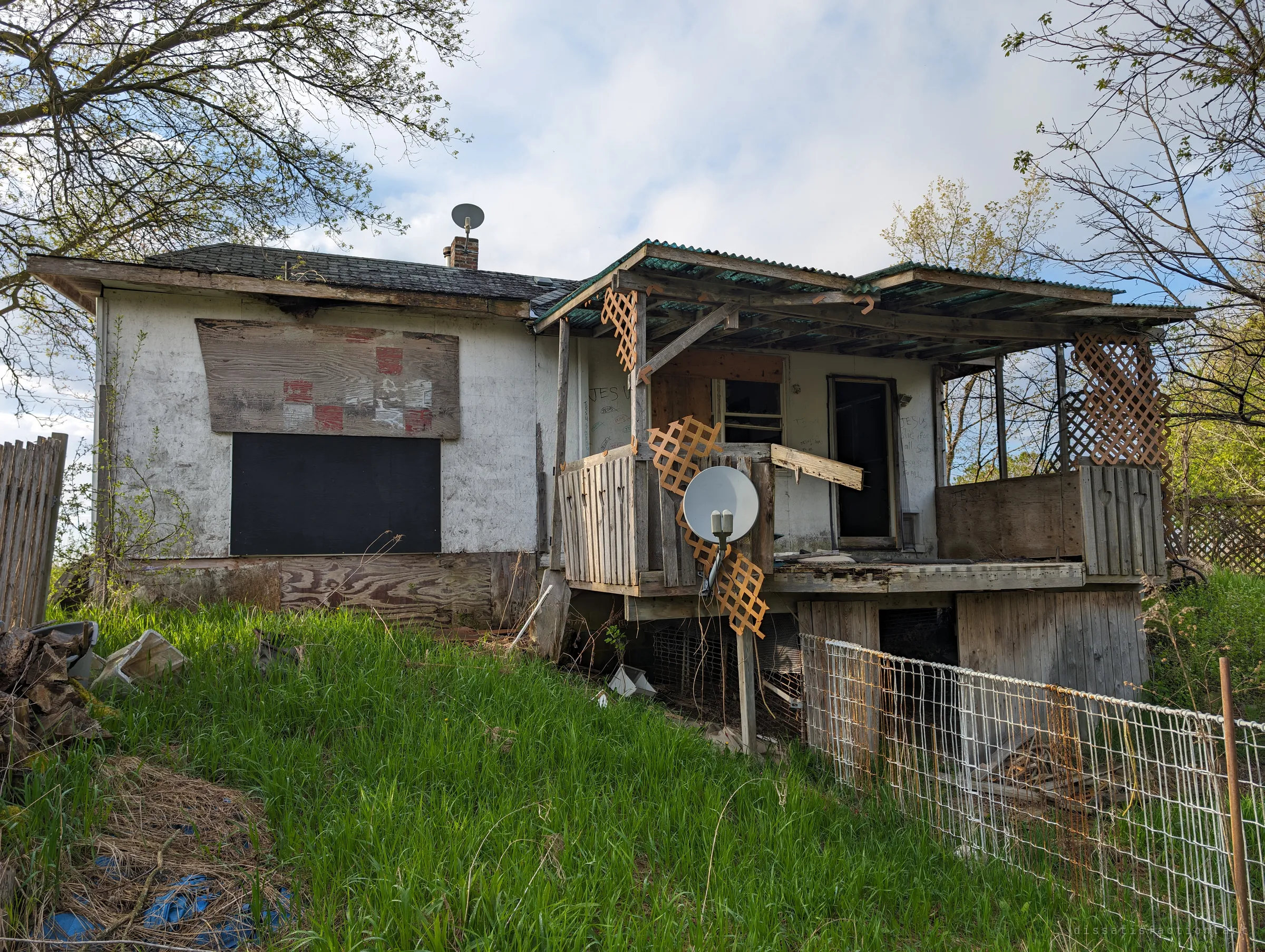 The back of the house, with boarded windows, a collapsing wooden deck wrapped in lattice and a satellite dish hanging off the railing. The rear shows how far the decay has gone.