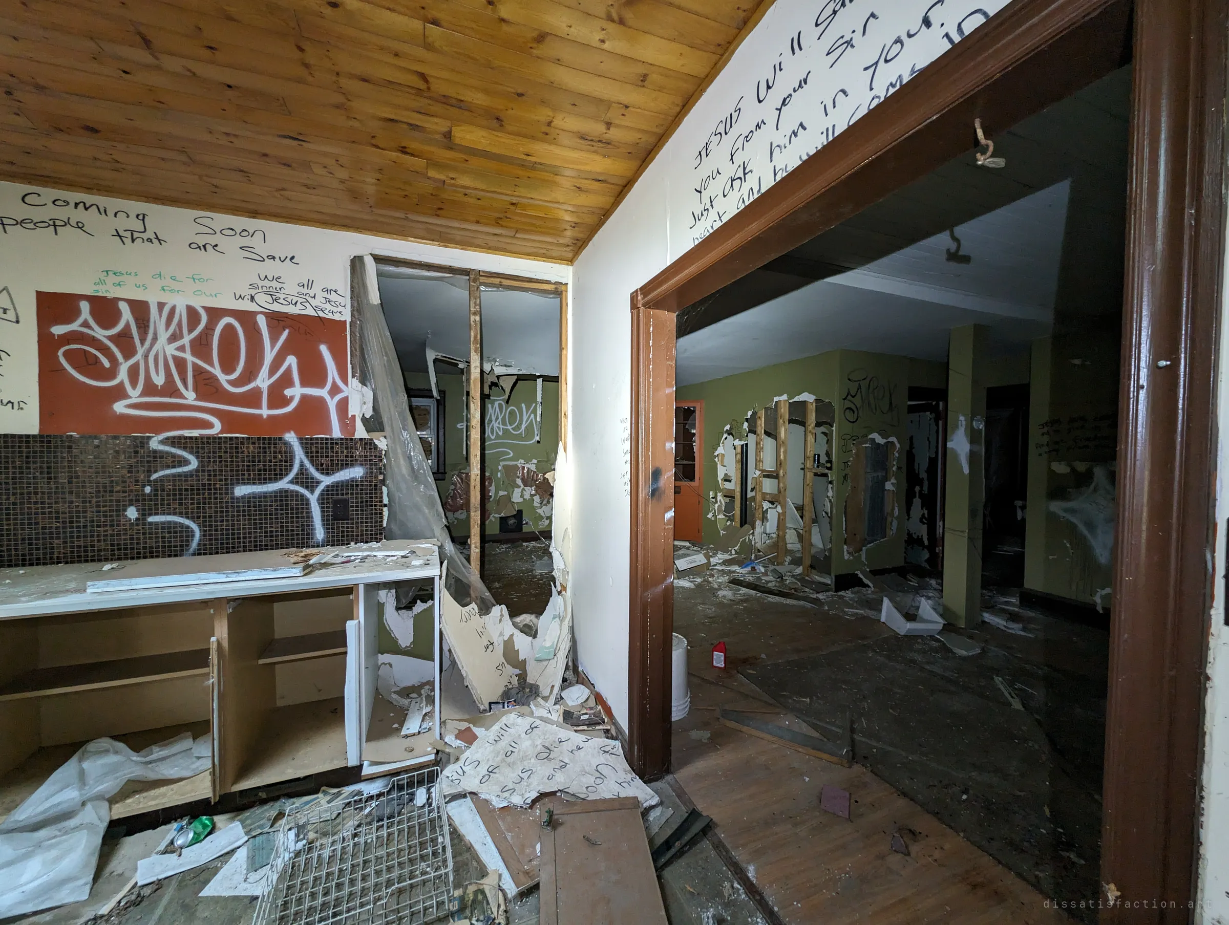 A gutted kitchen with a wood-plank ceiling opens into green rooms with walls torn down to the studs. Spray-paint tags sit beside hand-written religious messages covering the walls.