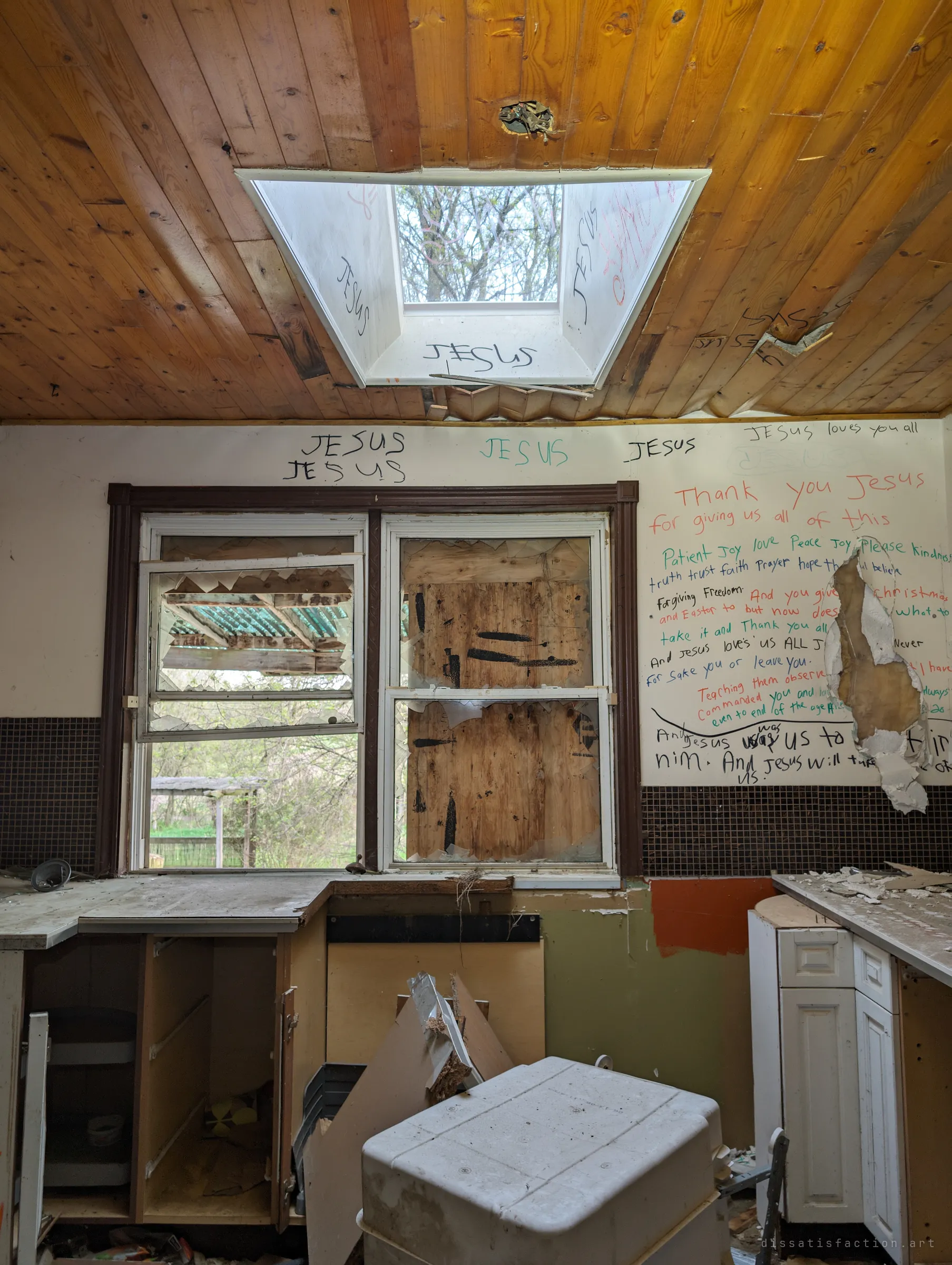 A skylight in the wooden kitchen ceiling above a half-boarded window, with religious messages written in coloured marker across the walls. Light from outside falls onto broken cabinets and a toppled freezer.