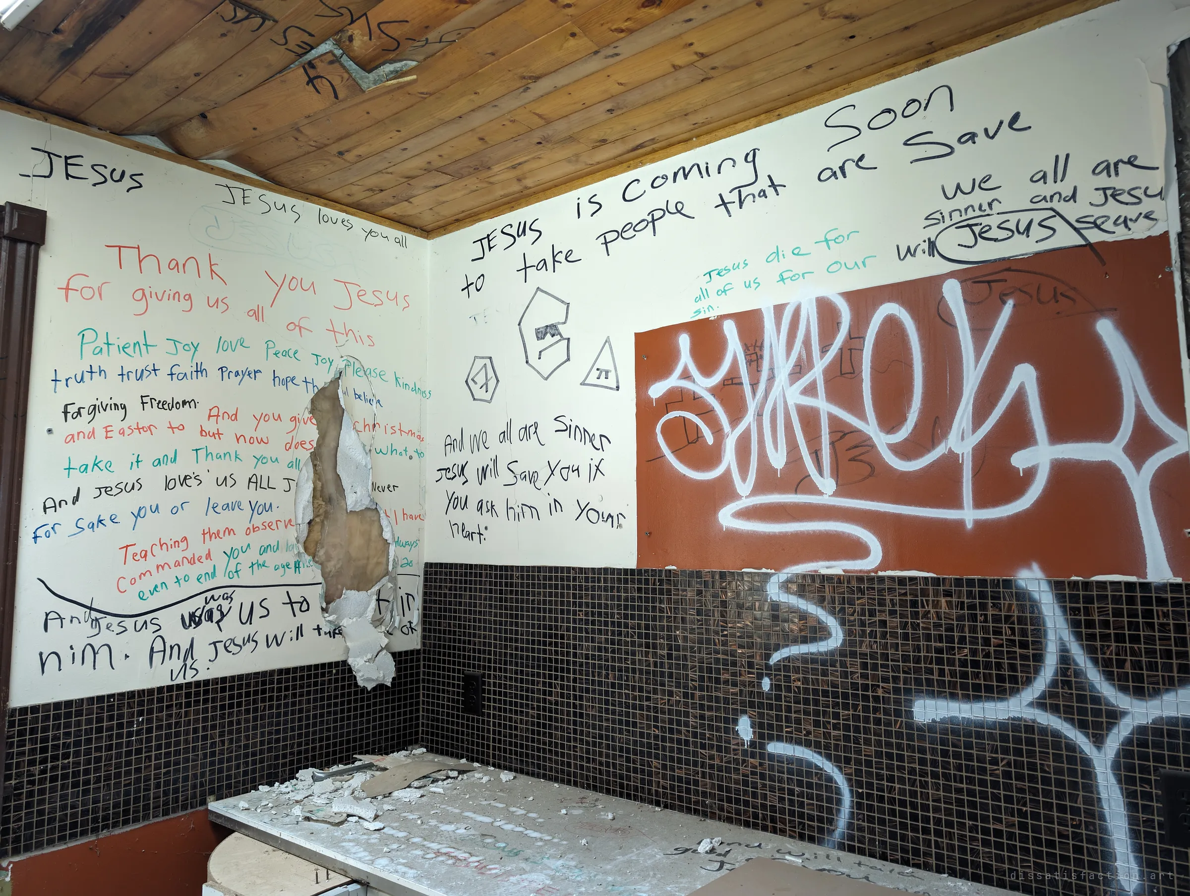 A kitchen corner where every bit of wall is covered in hand-written prayers and verses, next to a big white spray-paint tag over a tiled backsplash. The two kinds of writing sit side by side.