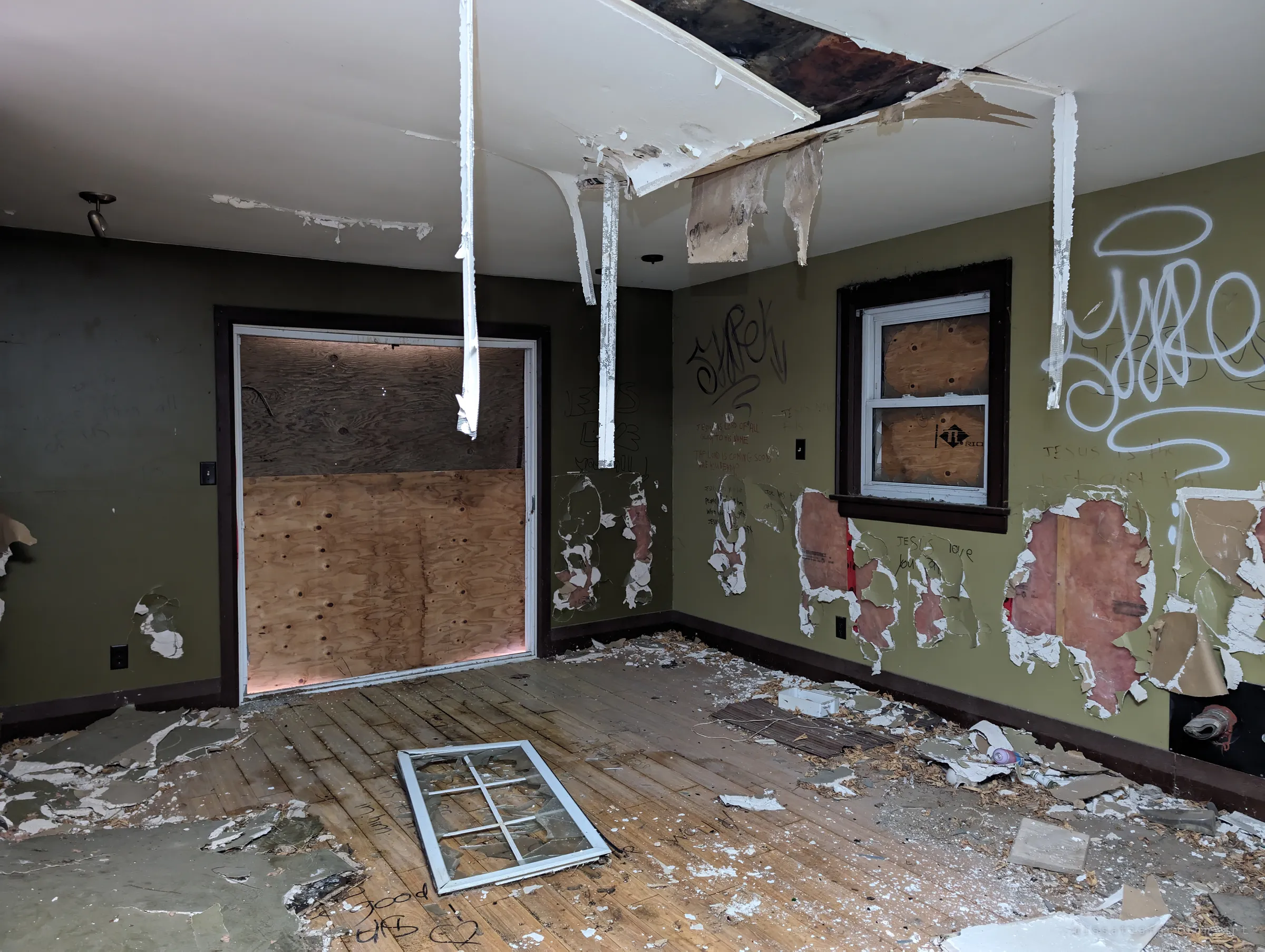 An olive-green living room with a plywood-covered patio door, drywall hanging in strips from the ceiling and a fallen window frame on the floor. Insulation shows through holes punched in the walls.