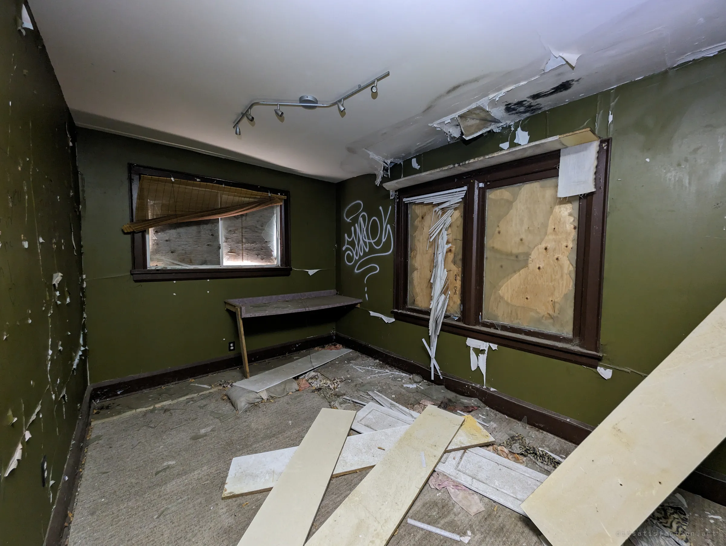 A small green bedroom with boarded windows, a track light still on the ceiling and loose boards scattered across the carpet. A white tag marks the corner wall.